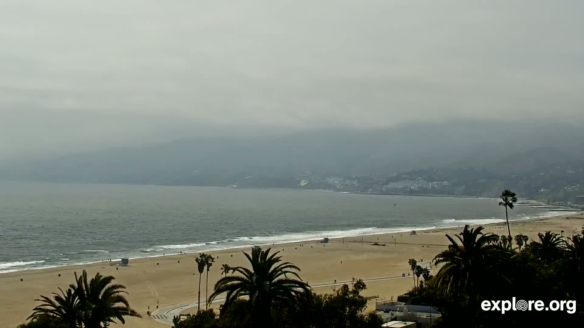 A hazy coastal scene shows a sandy beach with palm trees, a calm ocean with small waves, and distant, hazy mountains.