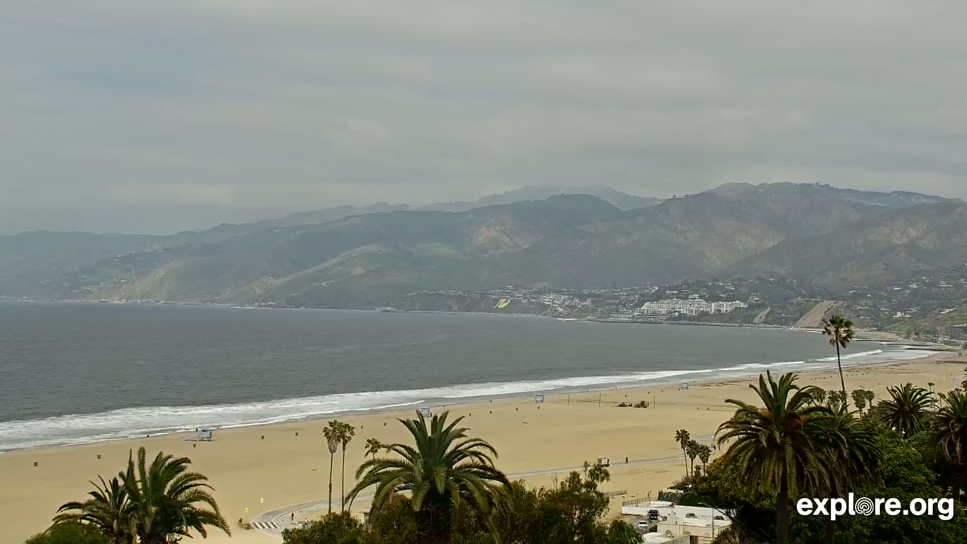 A sandy beach with palm trees and a calm ocean stretches out beneath a cloudy sky and a range of distant mountains.