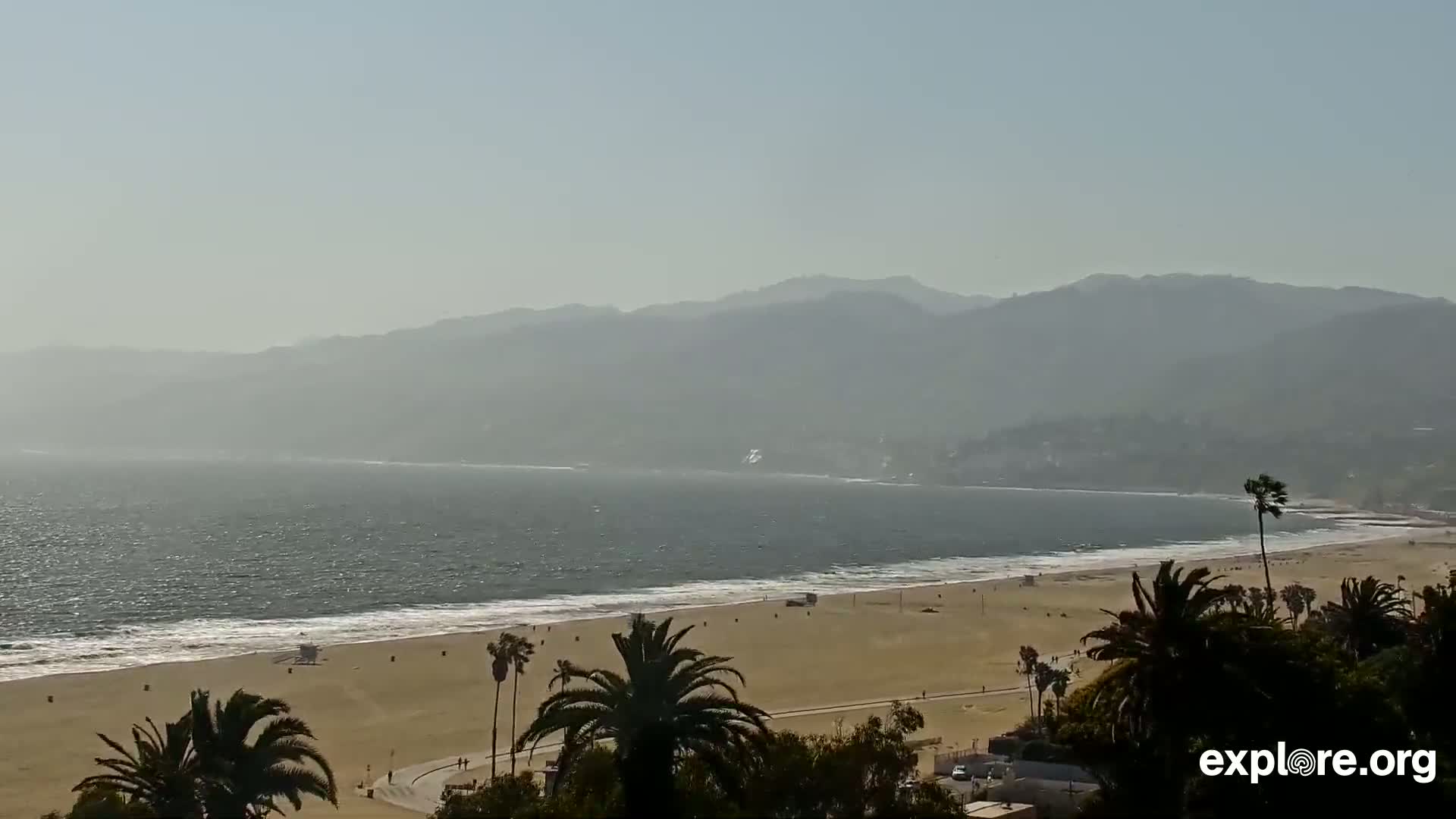 A hazy, sunny day reveals a sandy beach with palm trees, gentle waves lapping the shore, and distant mountains in the background.