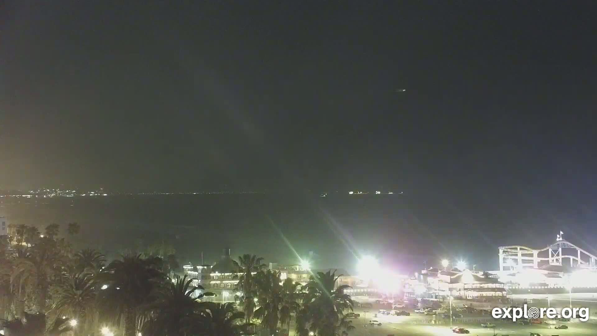 A nighttime view of a coastline shows a body of water, palm trees, and illuminated buildings including an amusement park ride.