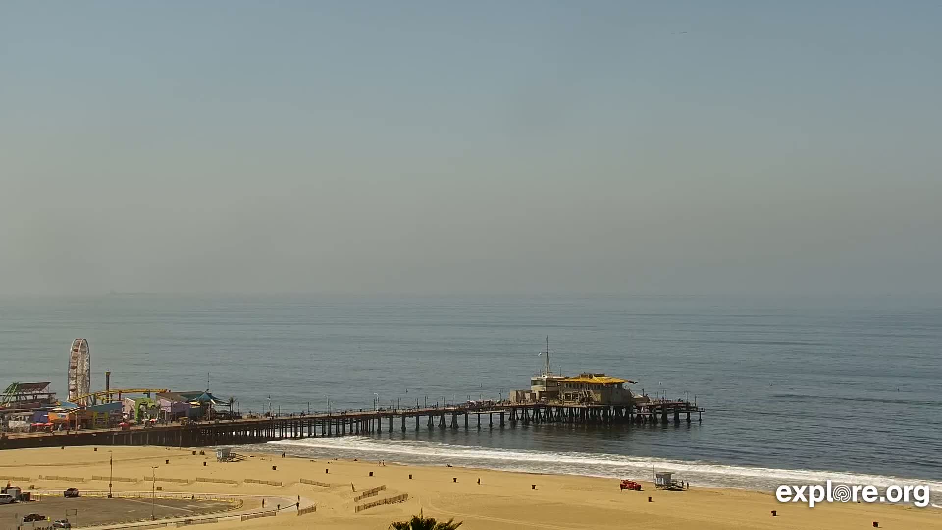 A long pier extends from a sandy beach into a hazy ocean, with a Ferris wheel visible in the background.