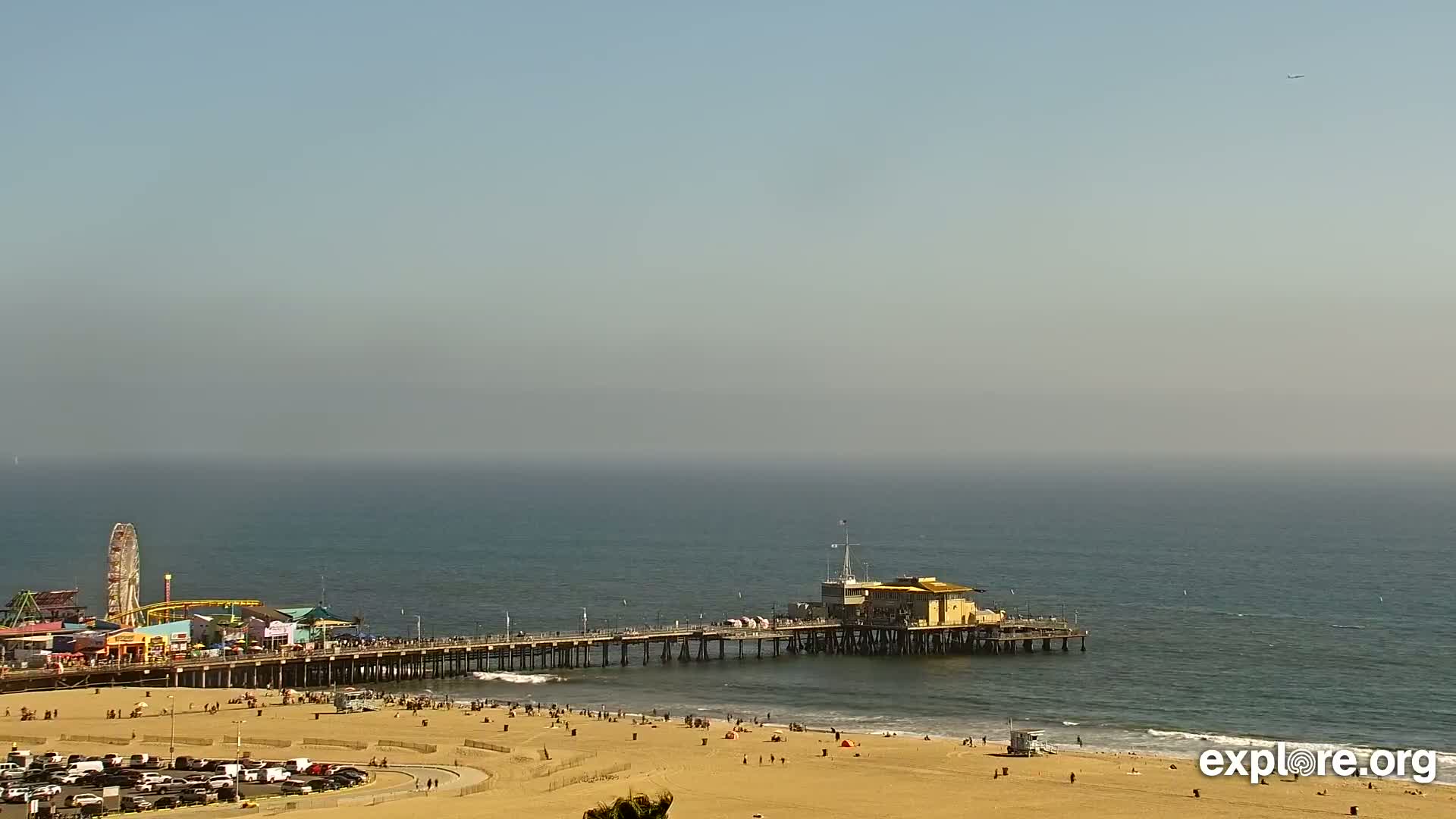 A long pier extends from a sandy beach into a hazy, calm ocean, with a Ferris wheel and other amusement park structures visible in the background.