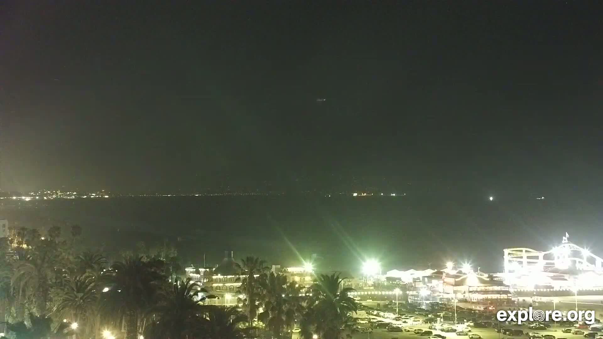 A nighttime coastal scene shows a body of dark water with city lights on the opposite shore, palm trees and brightly lit buildings and parking lots along a beachfront.