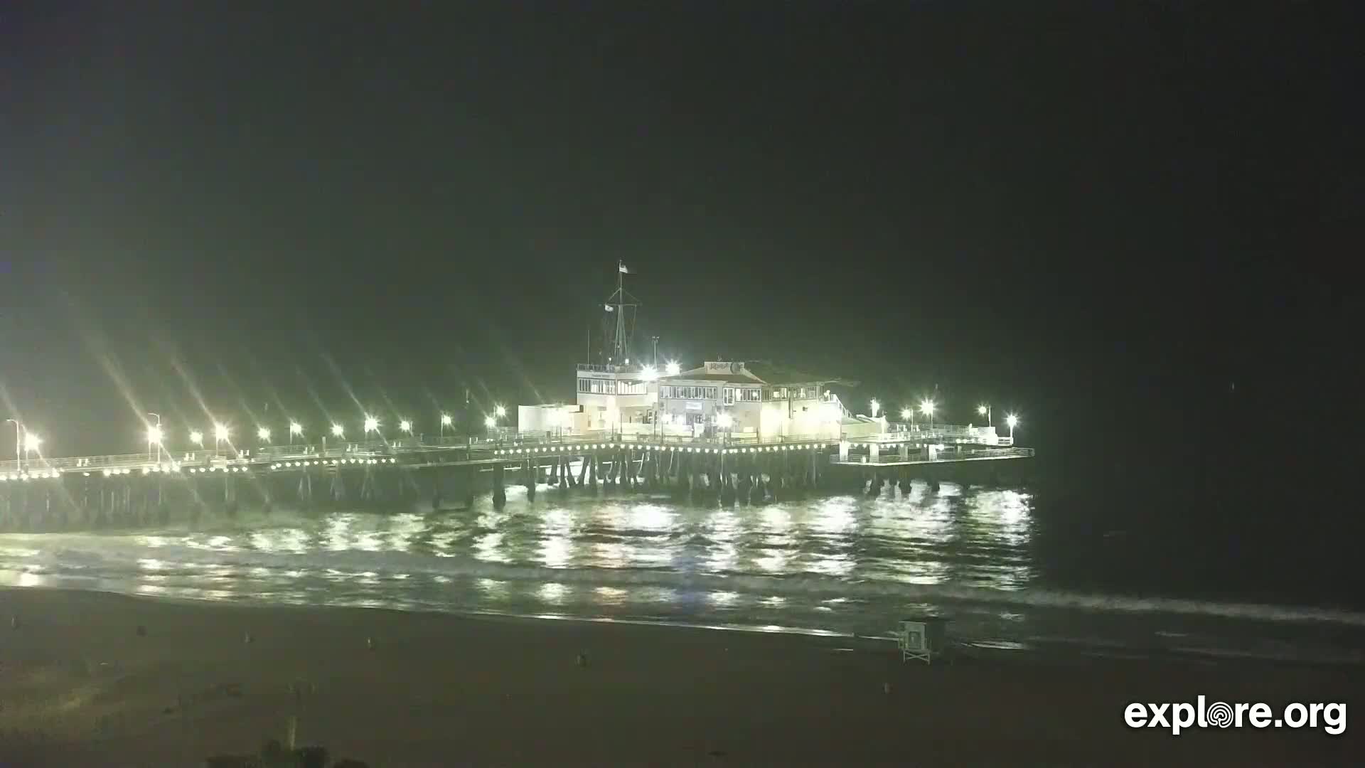 A brightly lit pier extends over a dark ocean with small waves under a night sky.