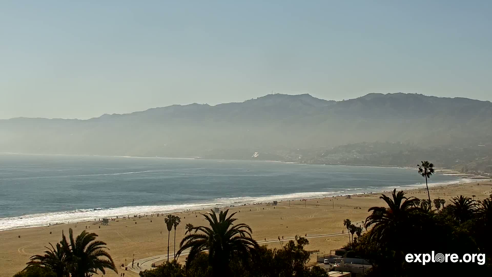 A hazy, sunny day reveals a sandy beach with palm trees, gentle waves lapping the shore, and a distant mountain range shrouded in mist.