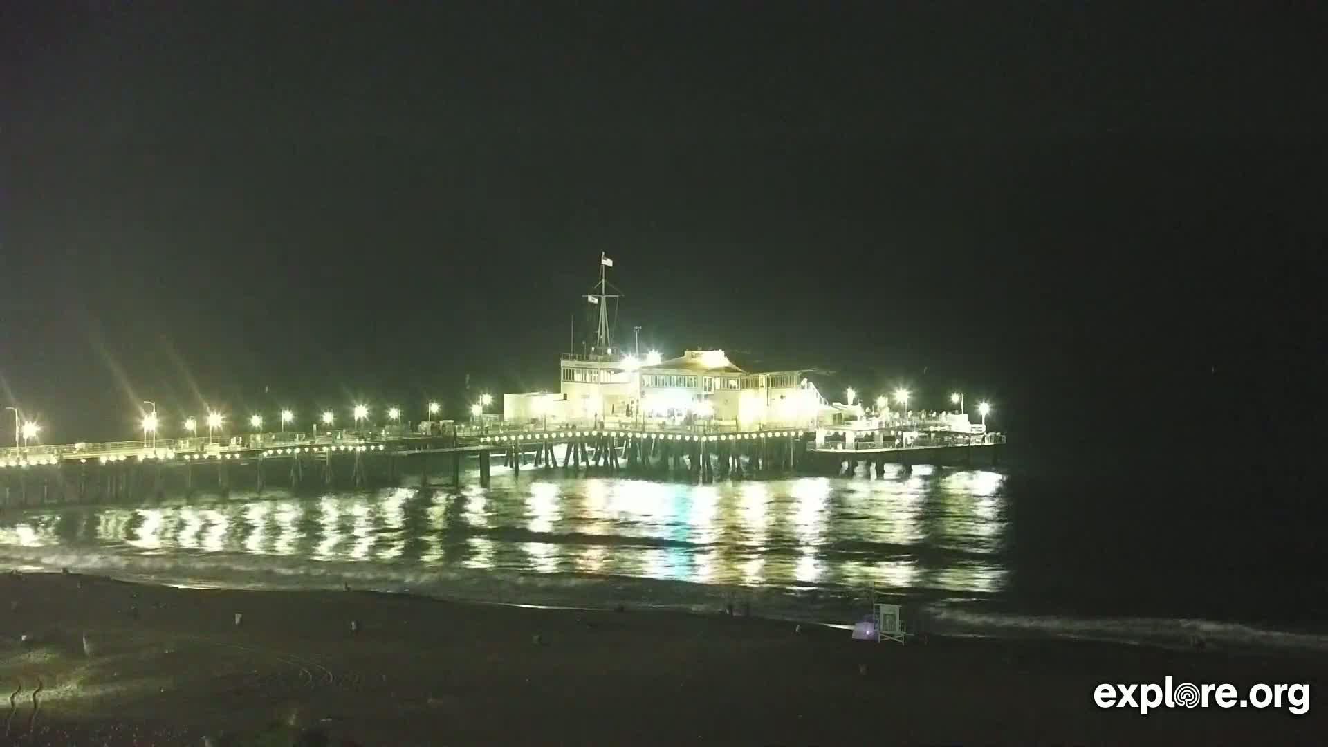 A brightly lit pier extends over a dark ocean at night, with gentle waves lapping the shore.