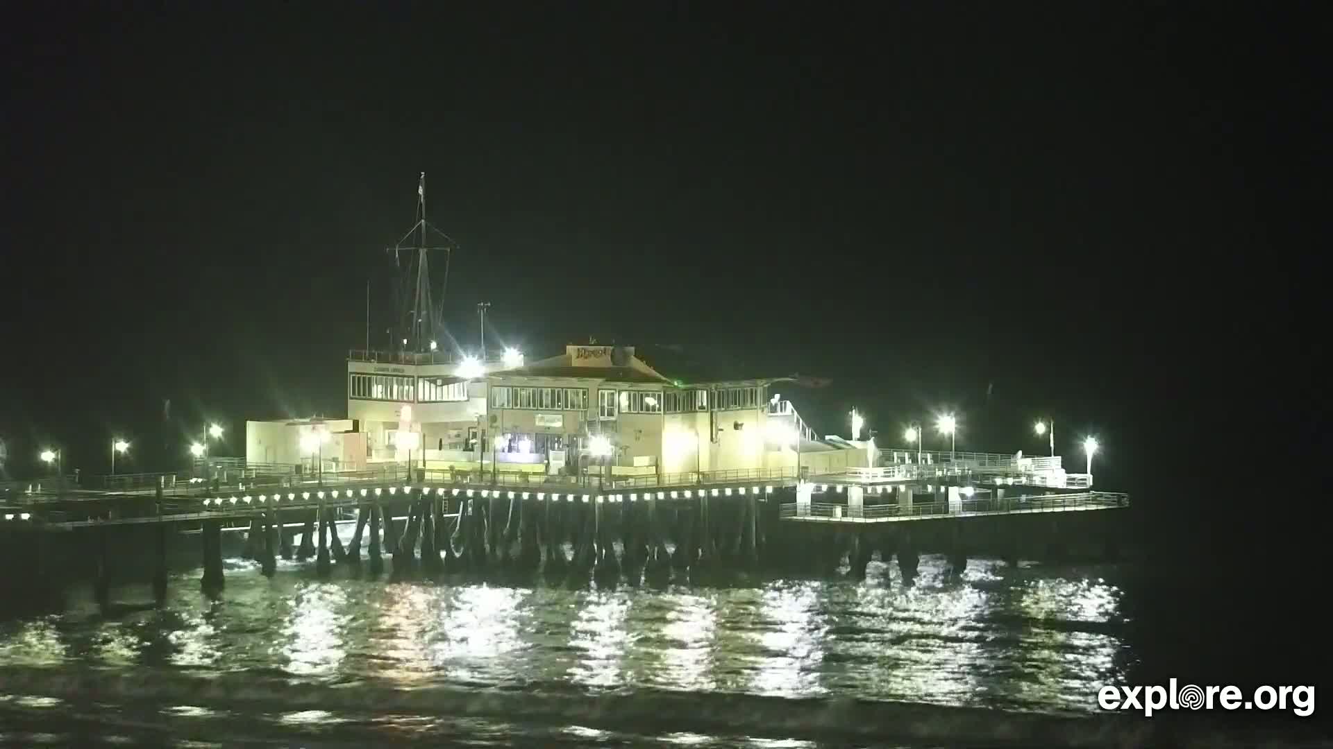 A brightly lit pier extends over dark, gently-lapping water at night.
