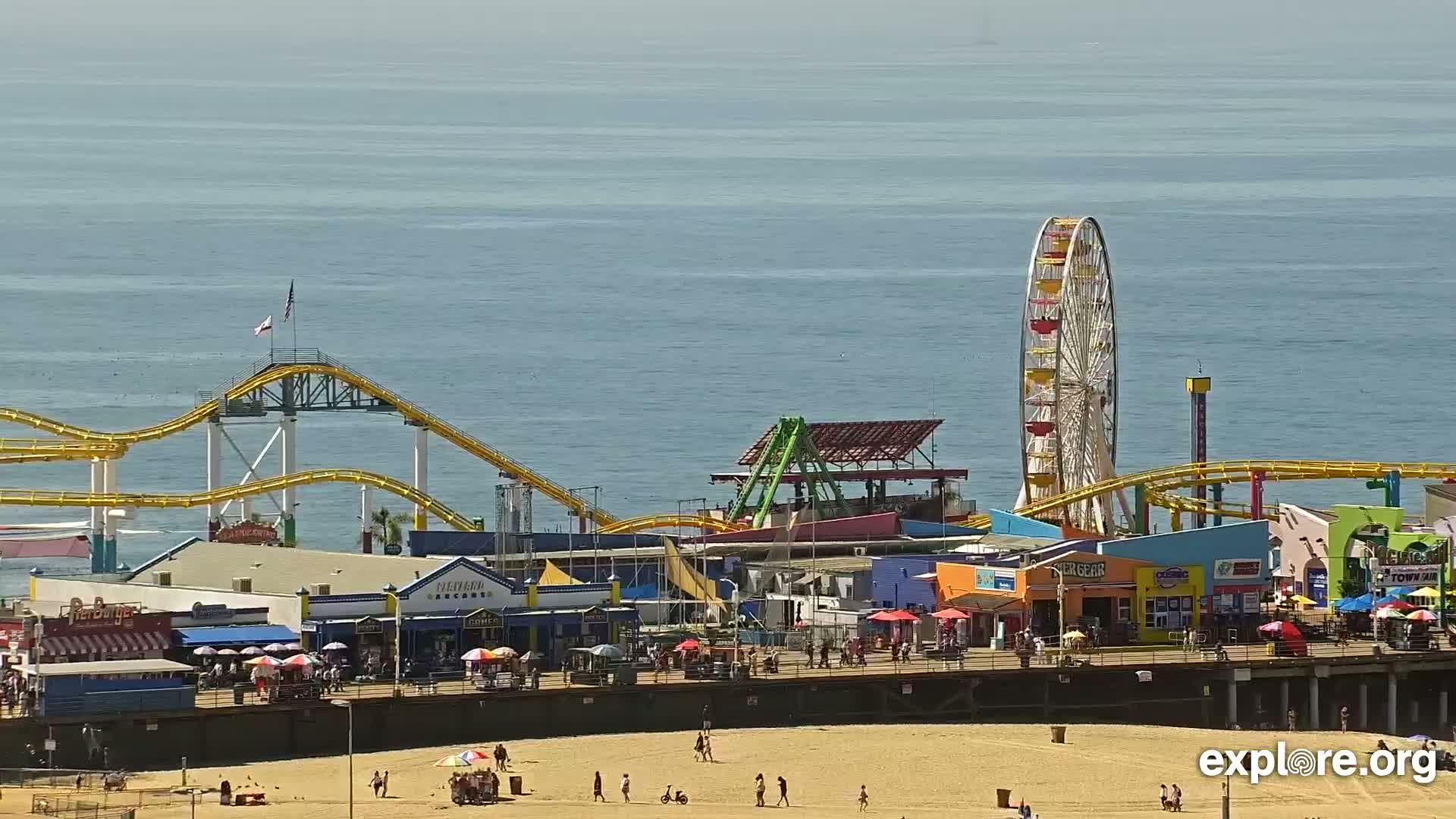 A brightly colored beachfront amusement park with a Ferris wheel and roller coaster is seen on a sunny day next to a calm ocean.