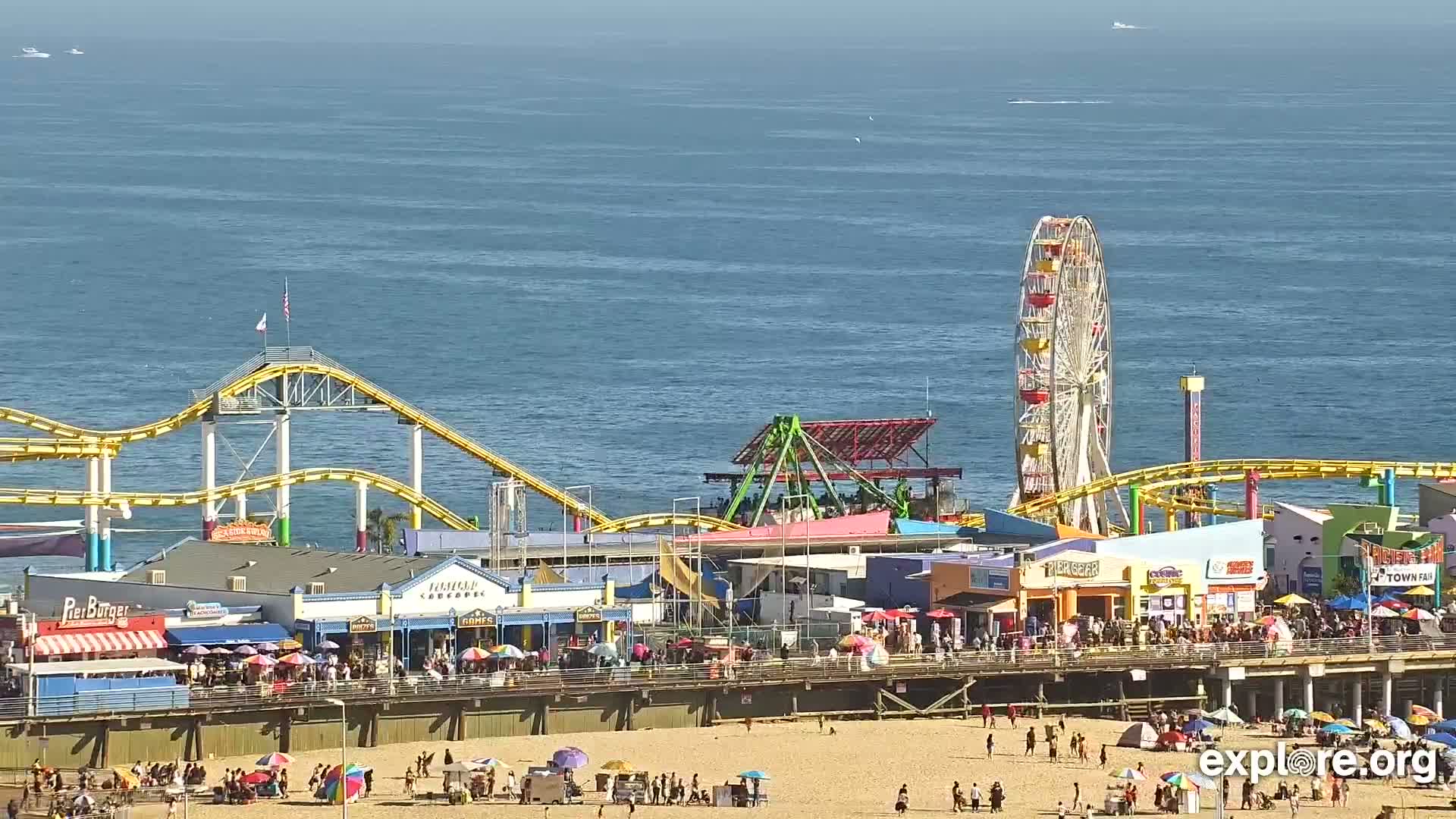 A brightly colored pier with a Ferris wheel, roller coaster, and numerous shops and restaurants overlooks a sandy beach and calm ocean on a sunny day.