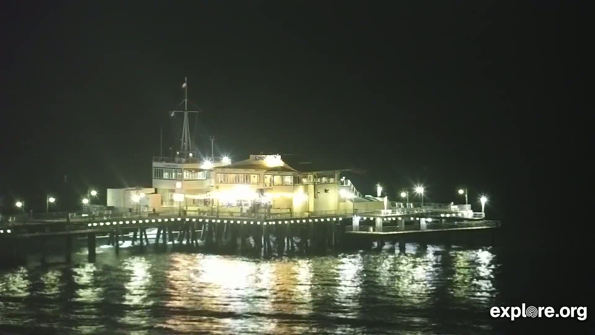 A brightly lit pier with a building on it stands in dark, calm water at night.