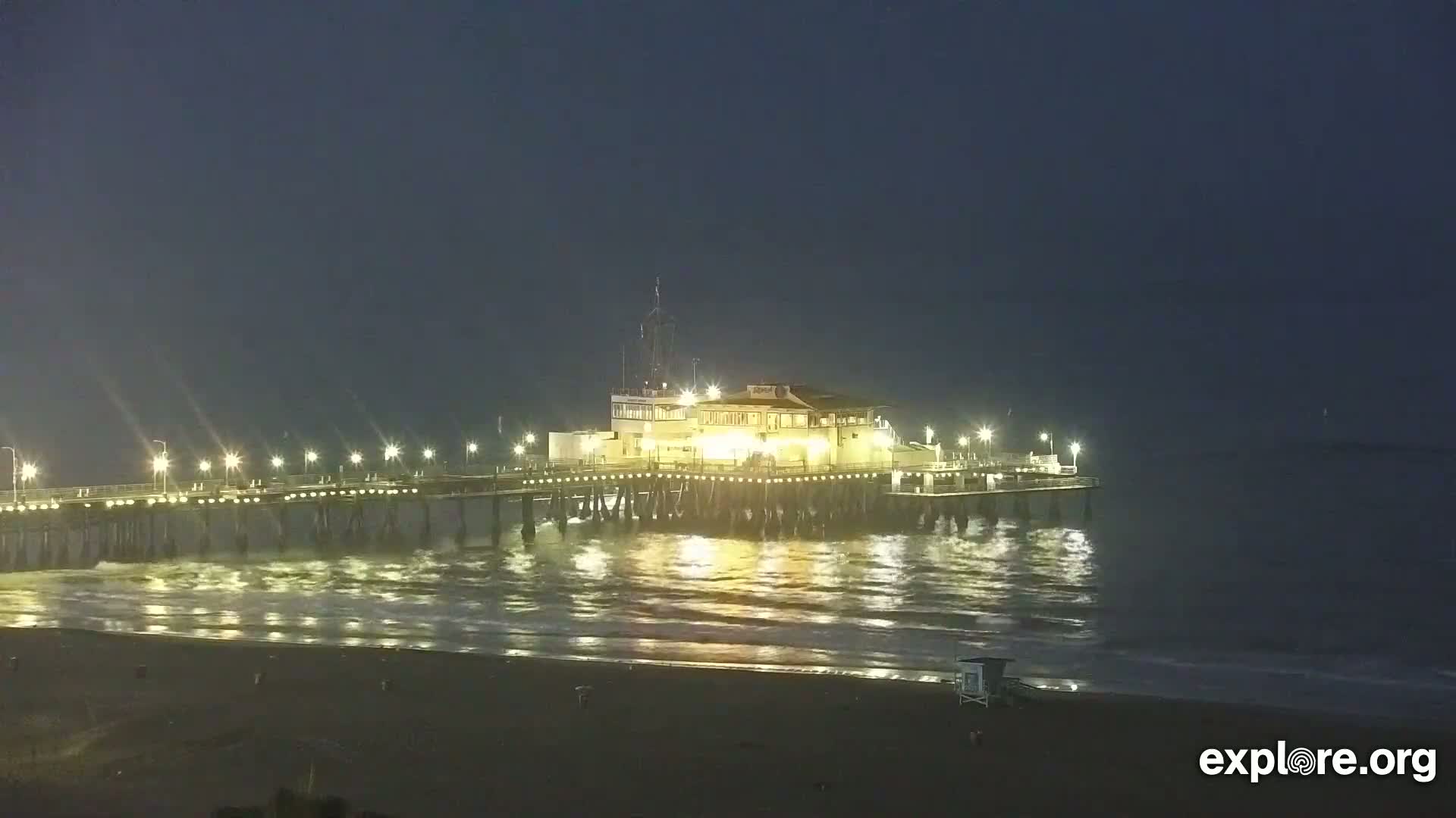 A brightly lit pier extends over dark ocean waves at night.