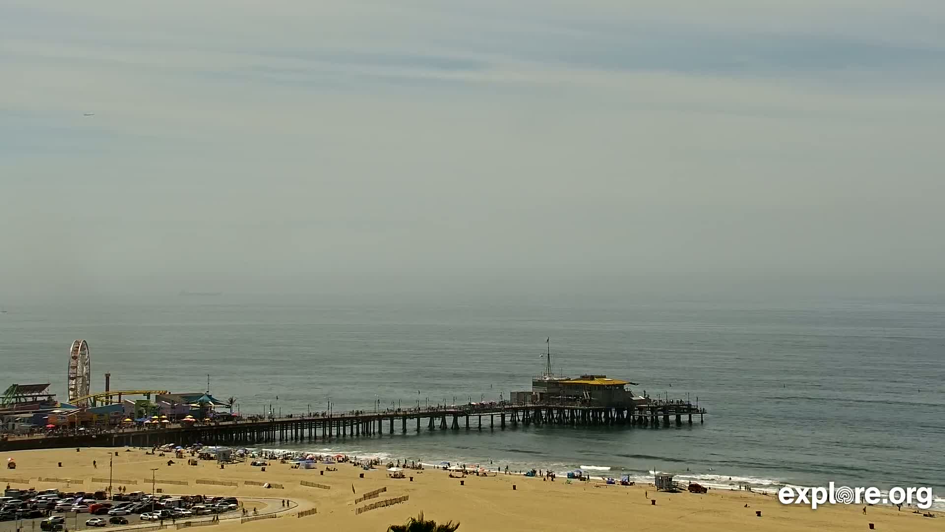 A hazy day shows a pier extending over a calm ocean, with a sandy beach below populated by people and parked cars.