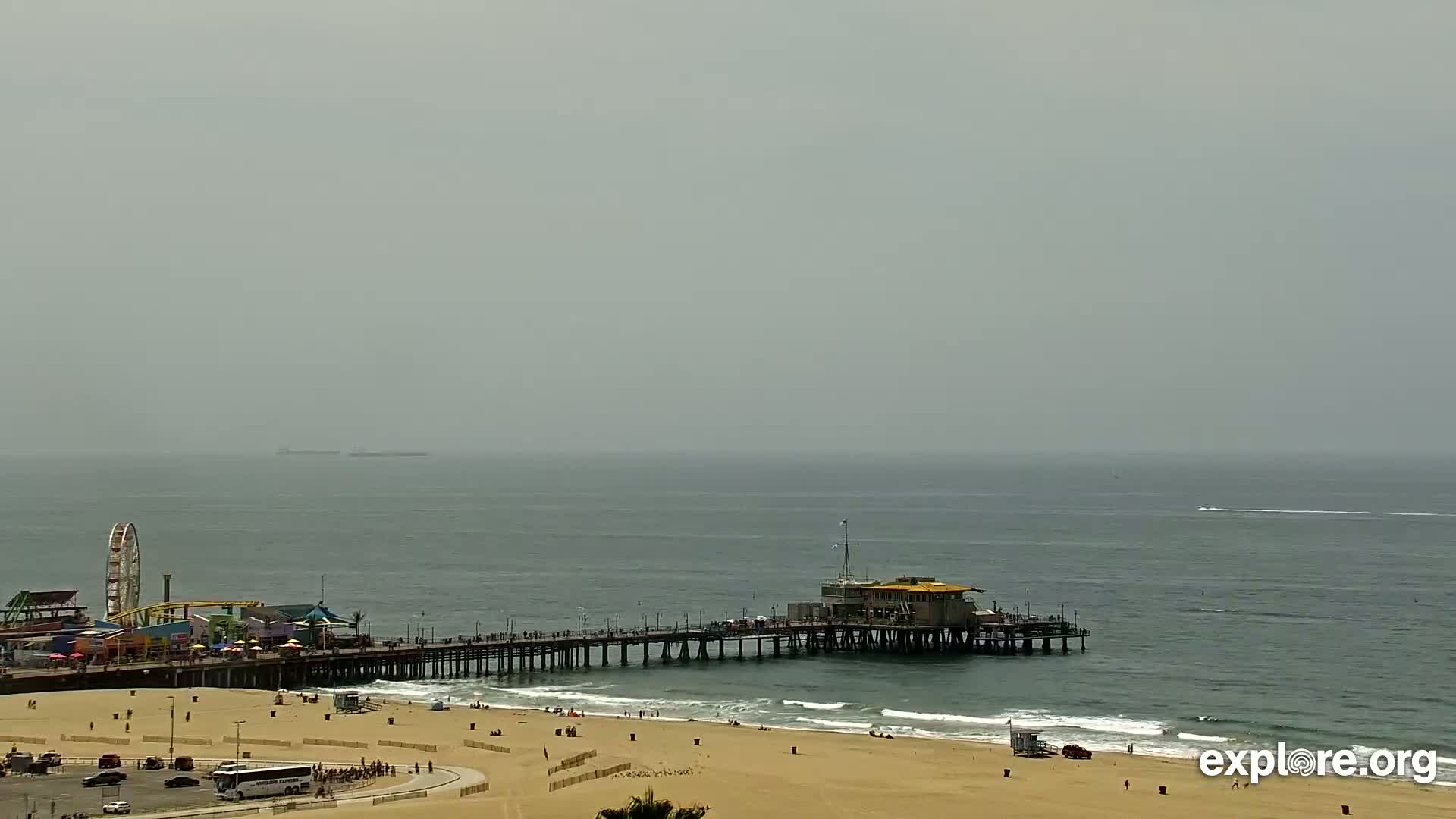 A long pier extends from a sandy beach into a hazy ocean, with a Ferris wheel and other amusement park structures visible on the shore.