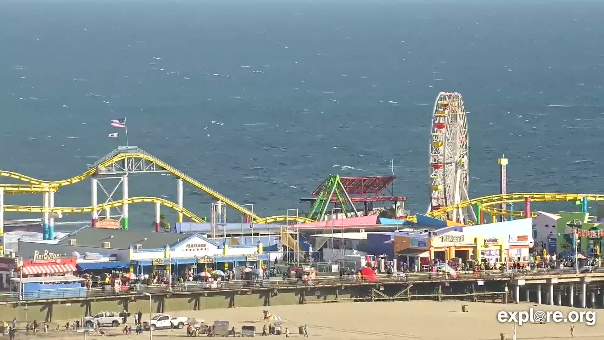 A brightly colored pier with a Ferris wheel, roller coaster, and various shops and restaurants sits on a sandy beach next to a somewhat choppy ocean under a sunny sky.
