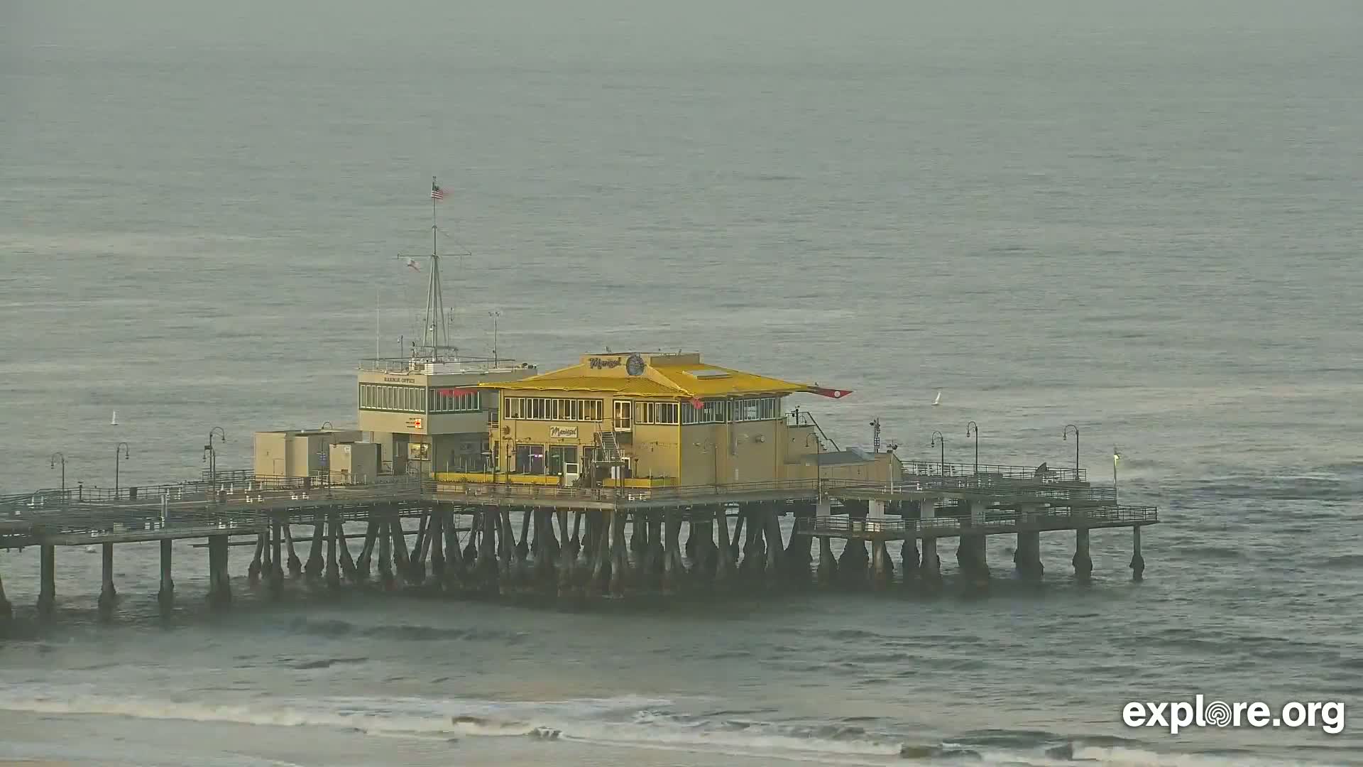 A long pier with a yellow building in the center extends over a choppy gray ocean under an overcast sky.