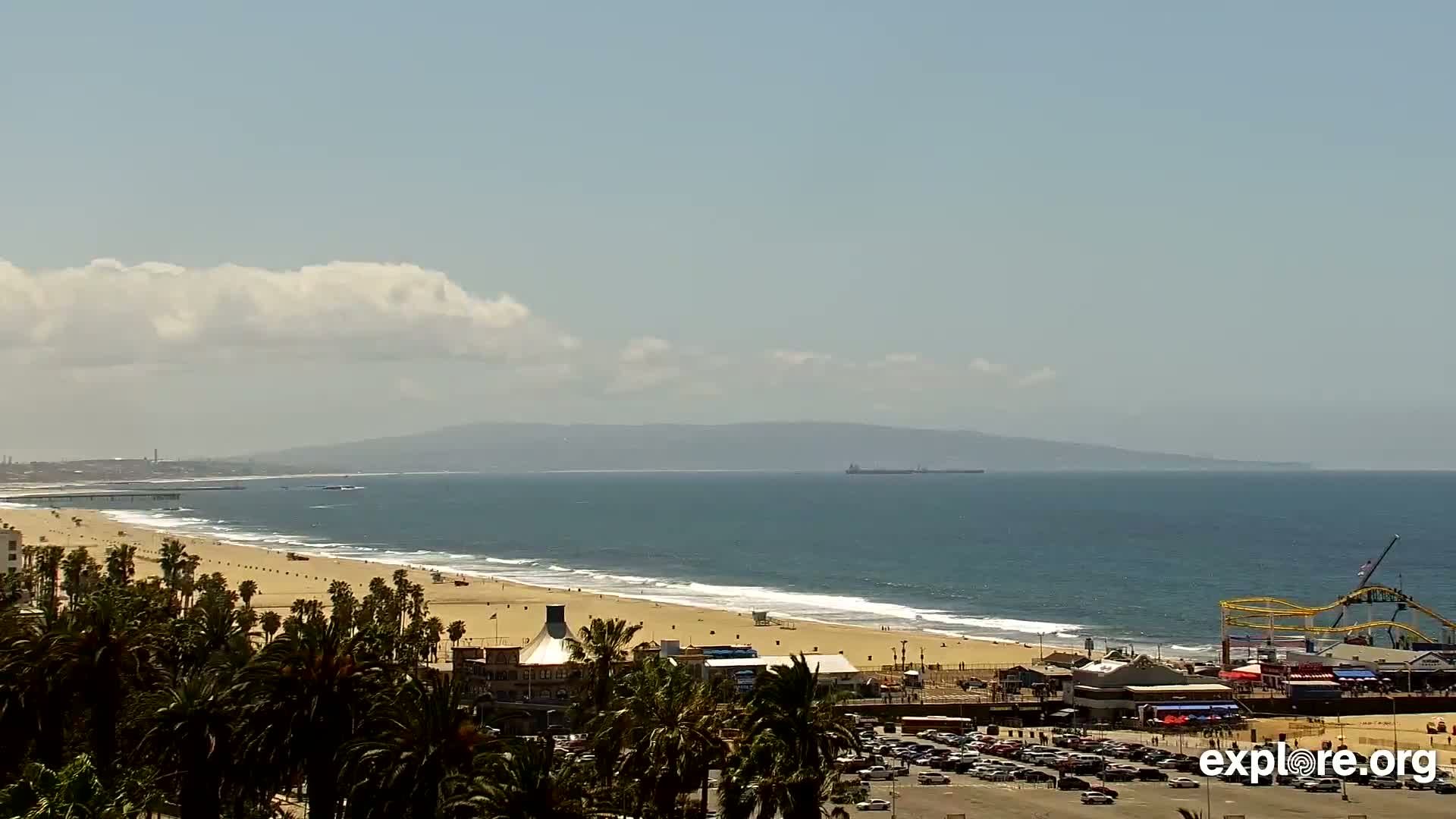 A sandy beach with gentle waves stretches along a coastline under a partly cloudy sky, backed by palm trees, buildings, and a roller coaster.