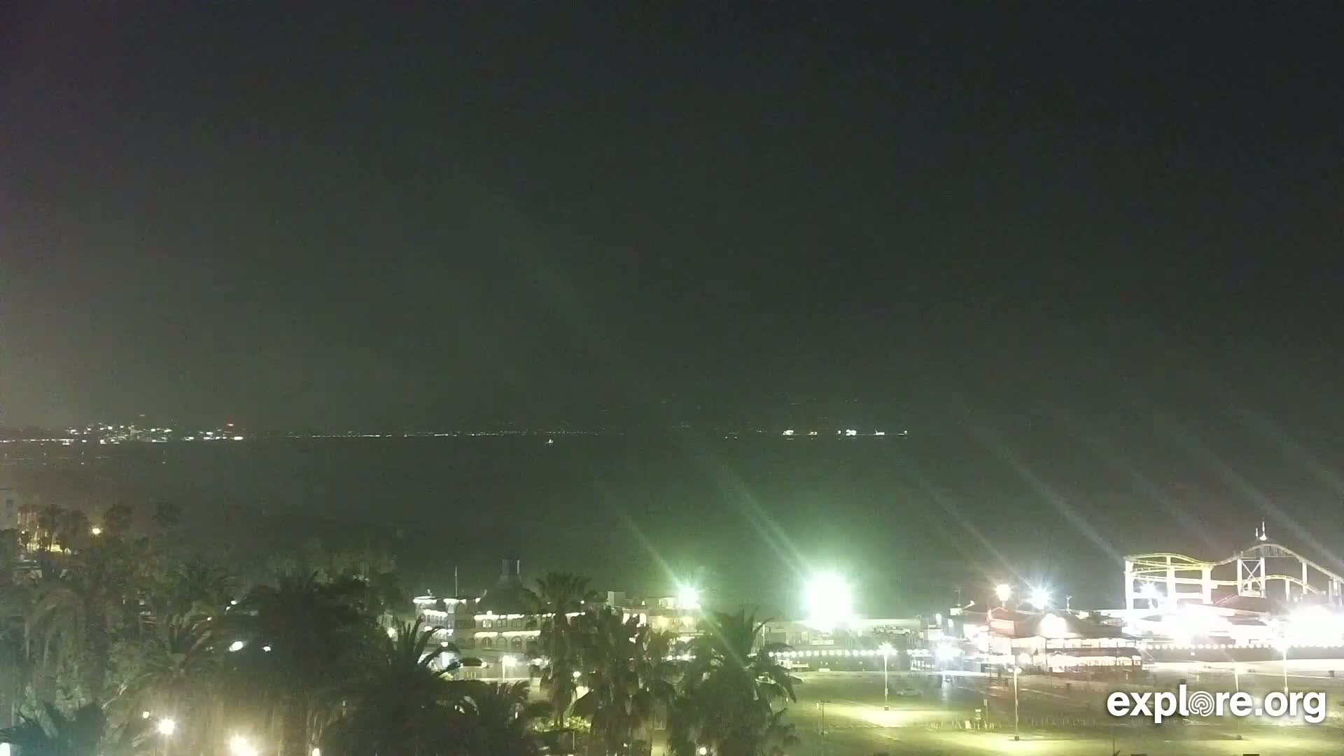 A nighttime view of a coastal city with palm trees, illuminated buildings, and a roller coaster in the distance over dark water under a clear sky.