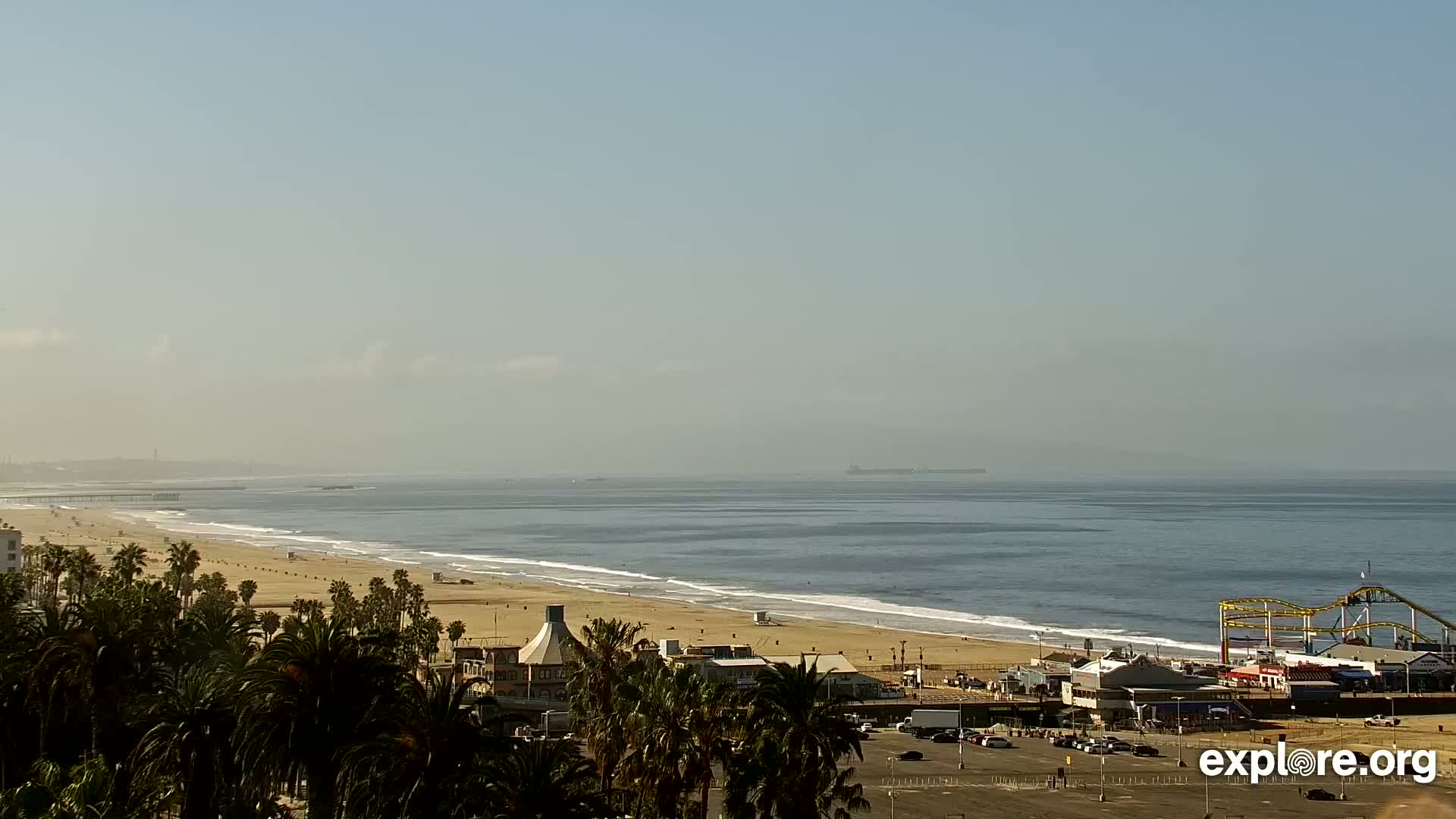 A hazy, sunny day reveals a long sandy beach with a pier and amusement park at the end, backed by palm trees and a parking lot.