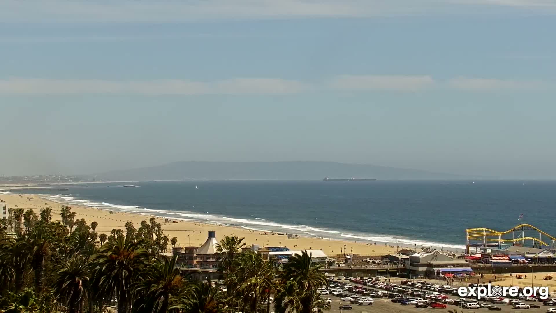 A long sandy beach with palm trees, a roller coaster, and a parking lot is visible beneath a hazy, sunny sky and a calm ocean with a distant cargo ship.