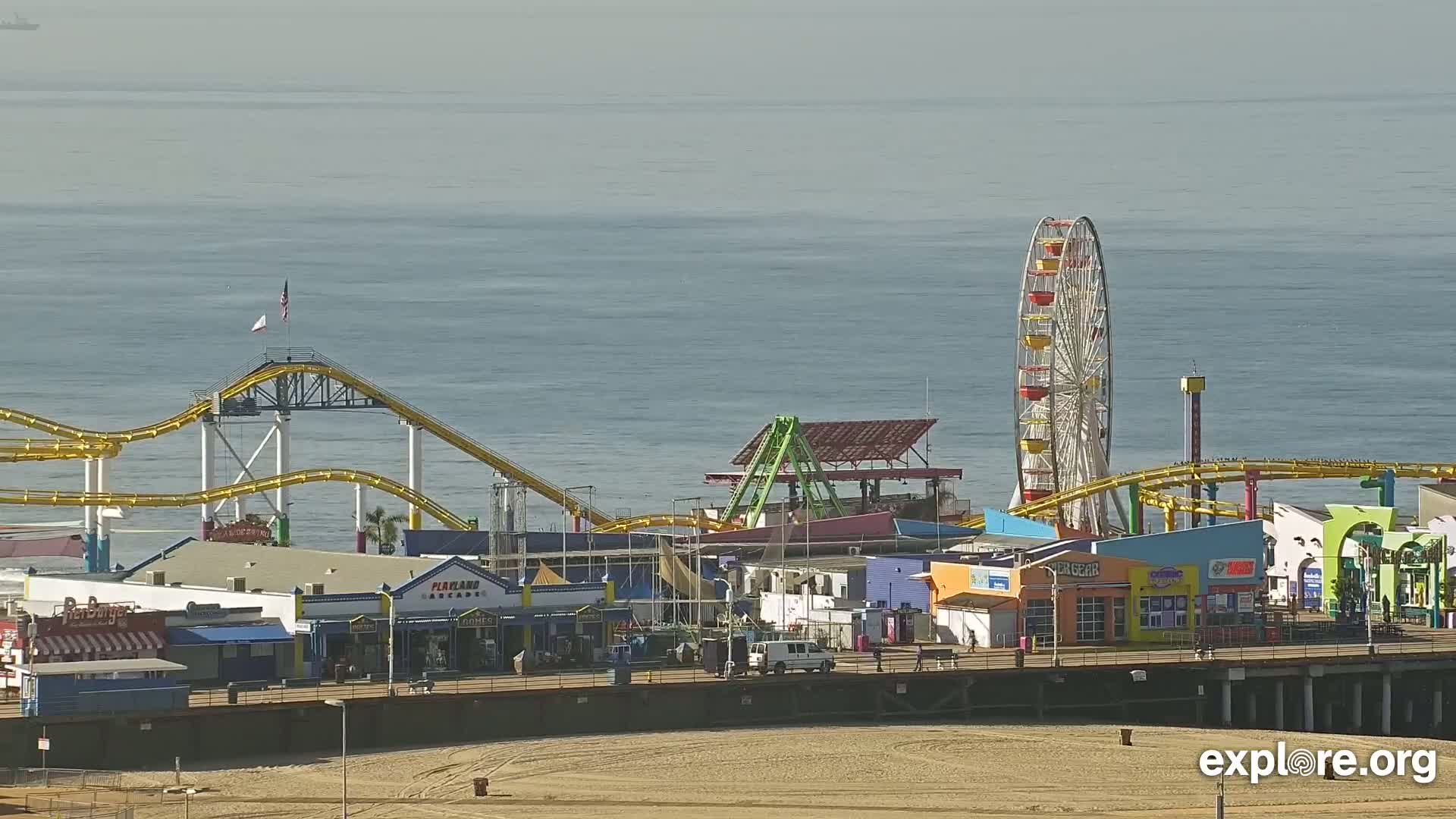 A brightly colored pier amusement park with a Ferris wheel and roller coasters is situated on a sandy beach next to calm ocean water under a hazy sky.