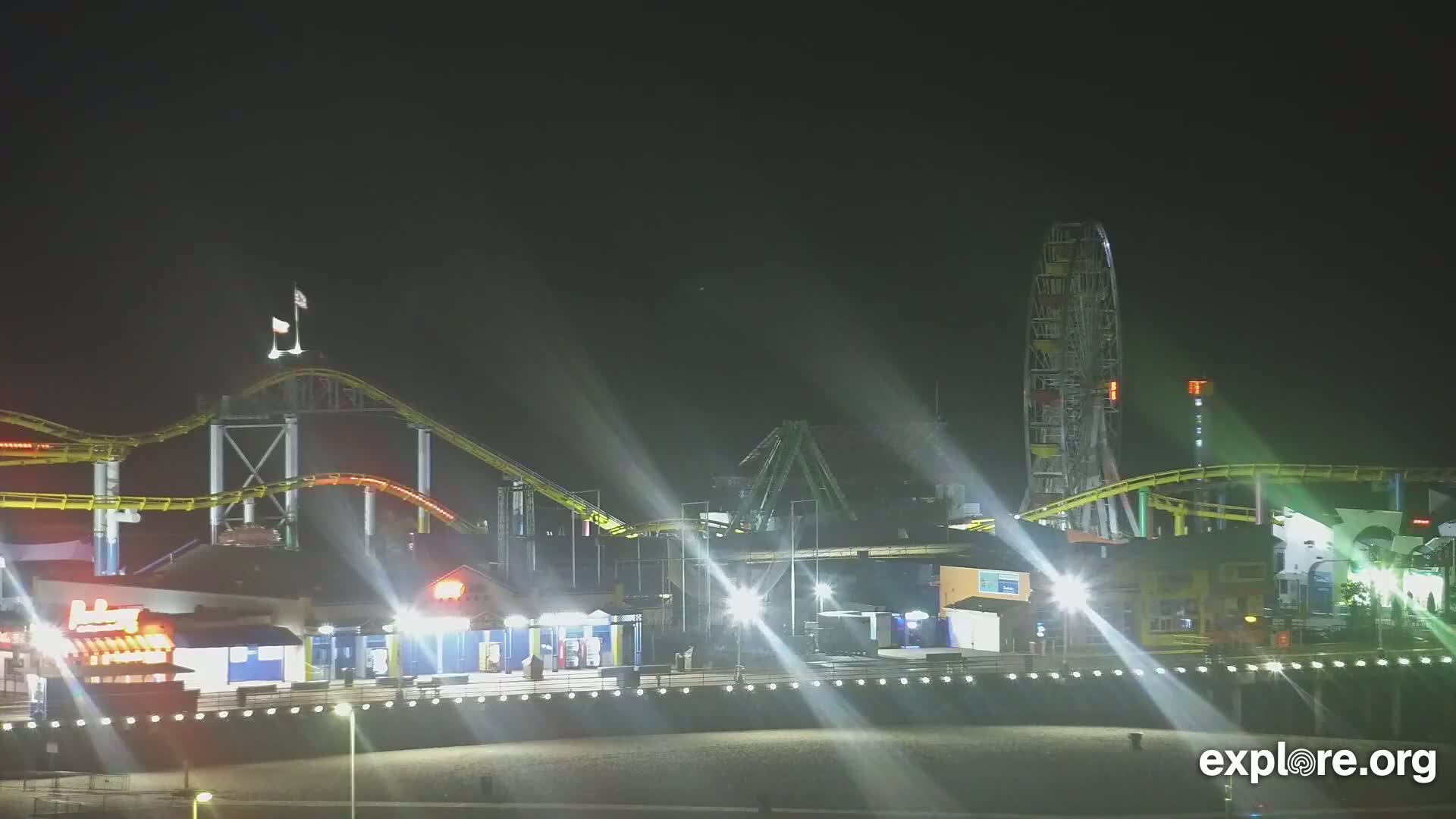 At night, brightly lit roller coasters and a Ferris wheel are visible at a beachfront amusement park.