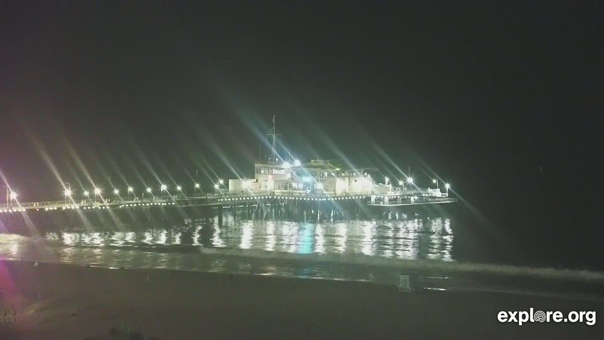 A brightly lit pier extends over a dark ocean with gentle waves under a clear night sky.