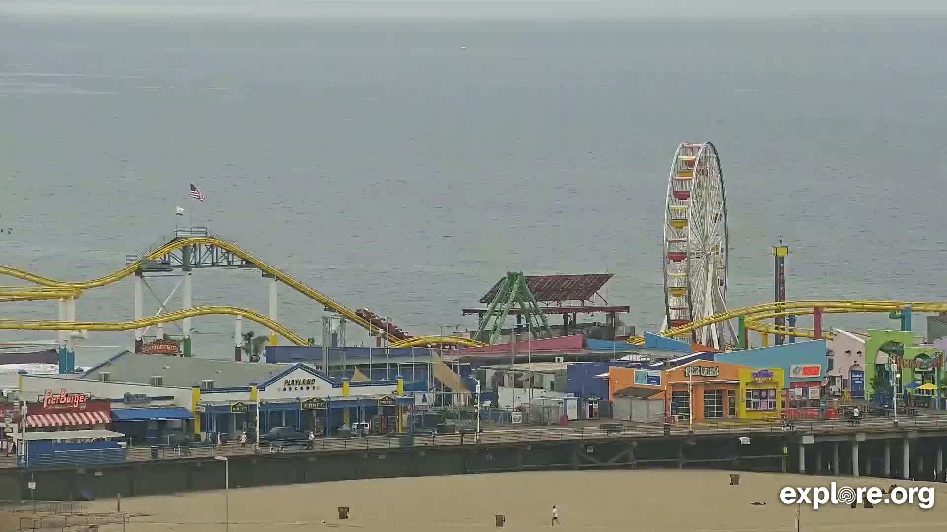 A pier with a Ferris wheel, roller coaster, and various shops and restaurants is seen on an overcast day next to a calm body of water.