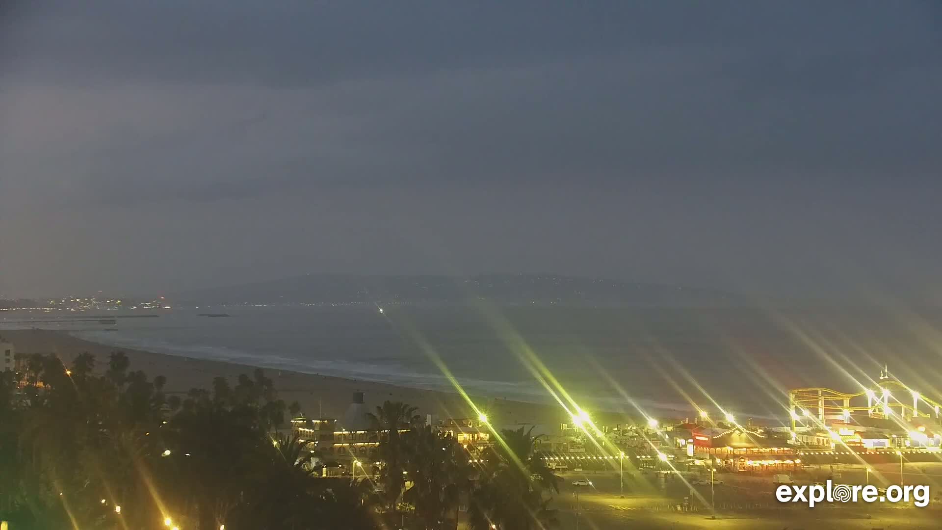 A nighttime view of a beach with a pier and roller coaster, under a hazy, dark sky.