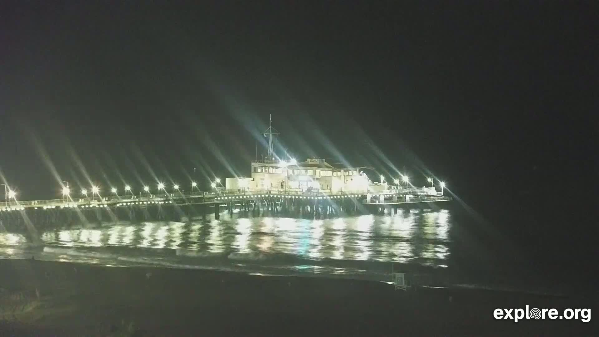 A brightly lit pier extends over dark, gently moving water at night.