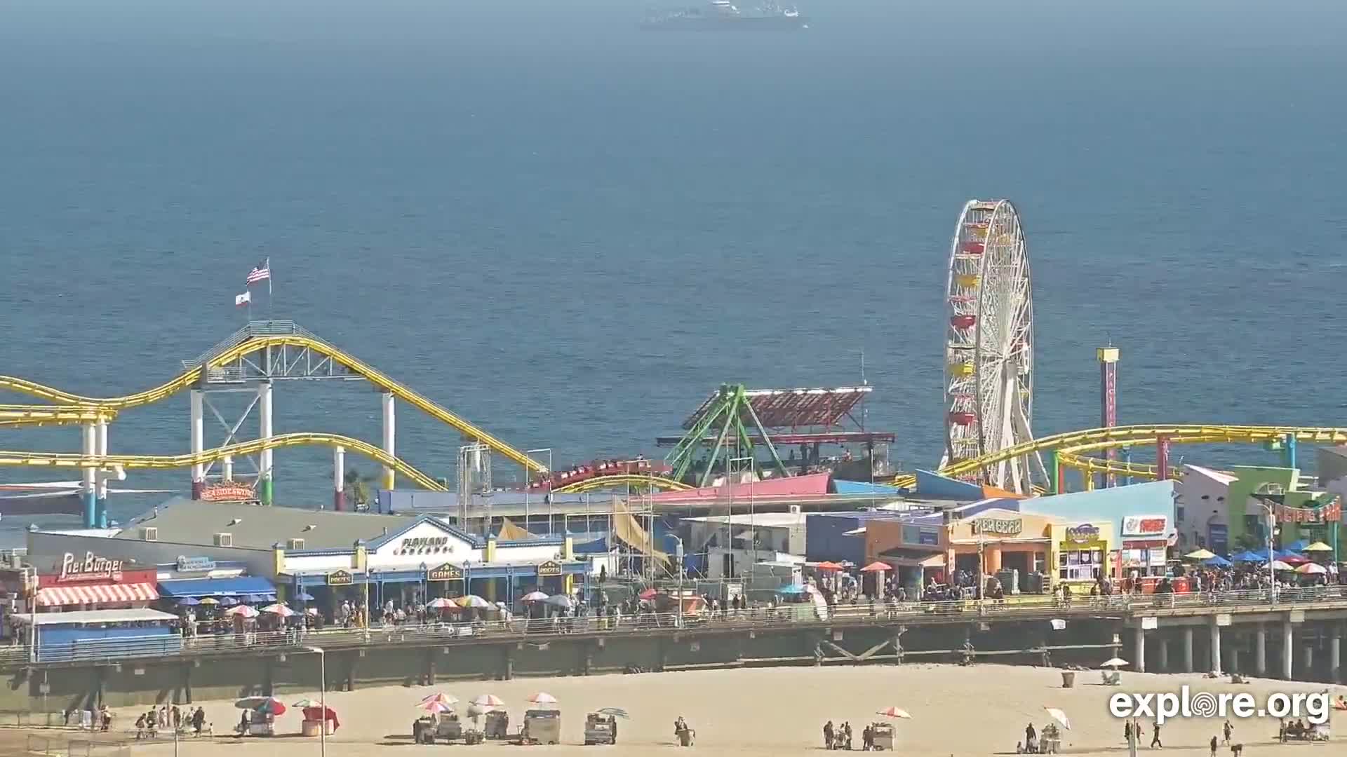 A pier with a Ferris wheel, roller coaster, and numerous shops and restaurants sits on a sandy beach next to a calm ocean.