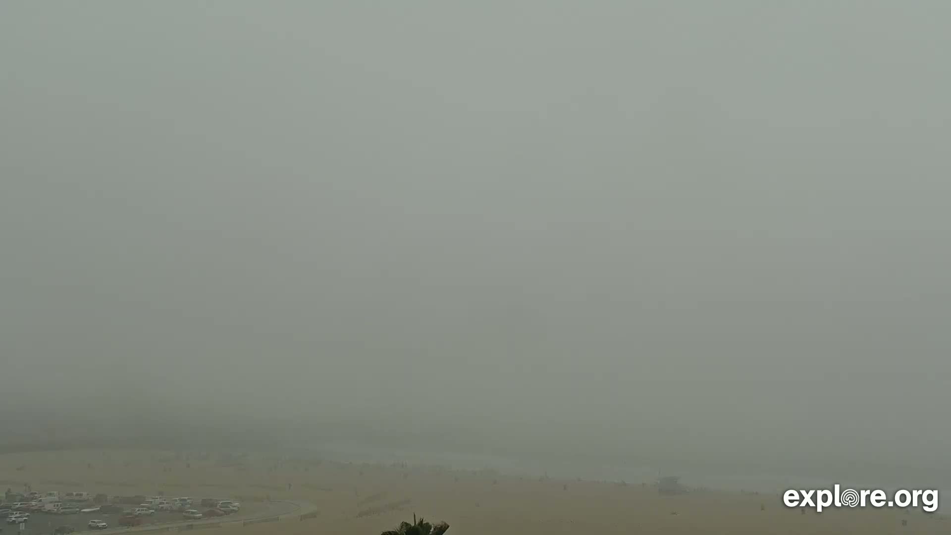 A hazy, foggy beach scene is visible, with a parking lot and some people sparsely scattered across the sand.