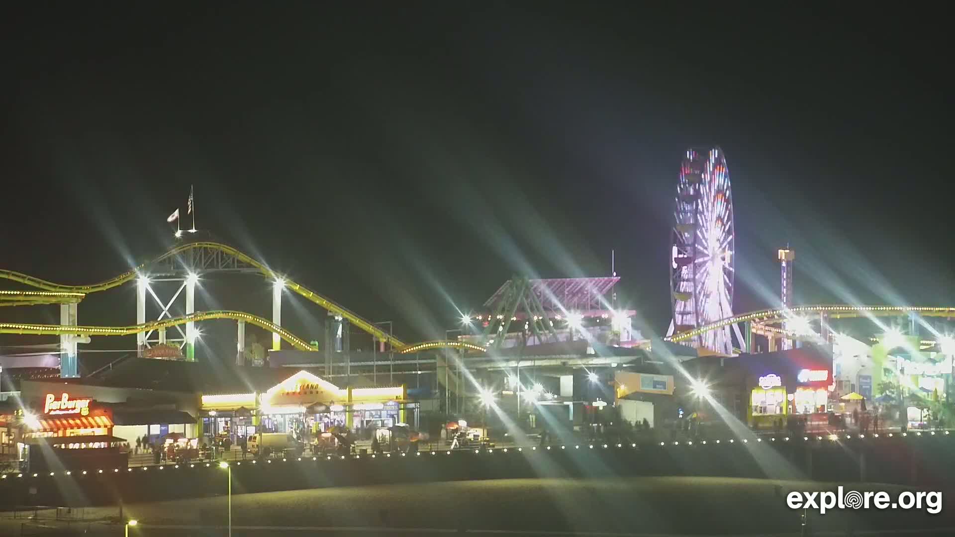 A brightly lit nighttime amusement park with a Ferris wheel and roller coaster, illuminated by strong light beams.