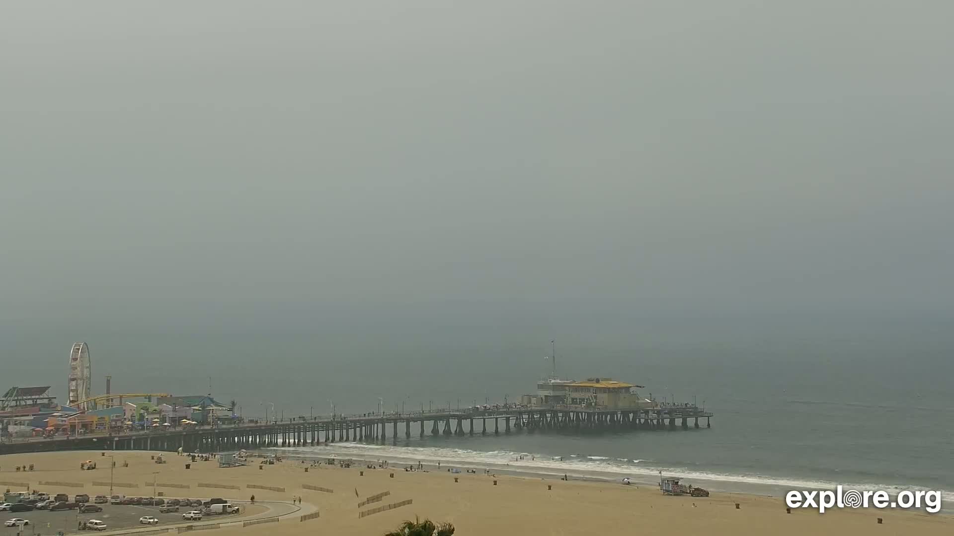 A hazy, overcast day reveals a pier extending into a calm ocean, with a beach and amusement park visible on the shore.