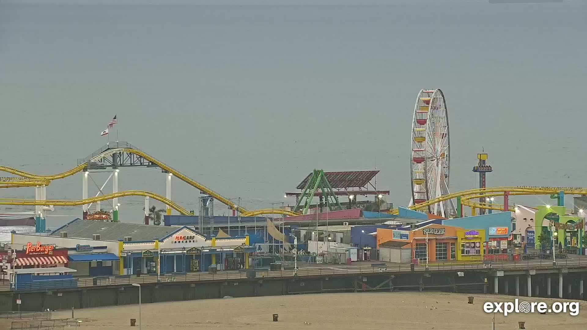 A brightly colored pier with roller coasters, a Ferris wheel, and various shops sits on a sandy beach overlooking a calm, overcast ocean.