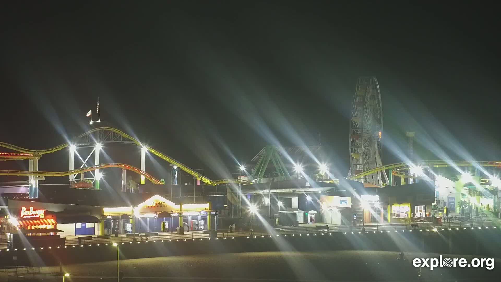 A brightly lit amusement park at night, featuring a roller coaster and Ferris wheel, is seen under a dark, clear sky.