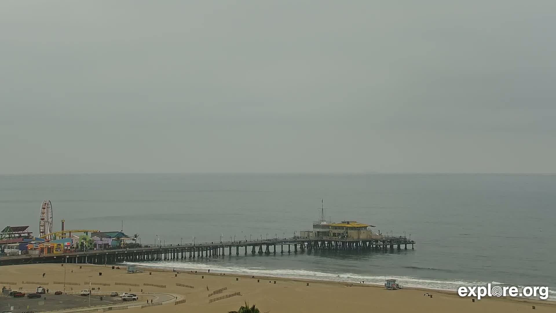 A long pier extends from a sandy beach into a calm, gray ocean under an overcast sky.