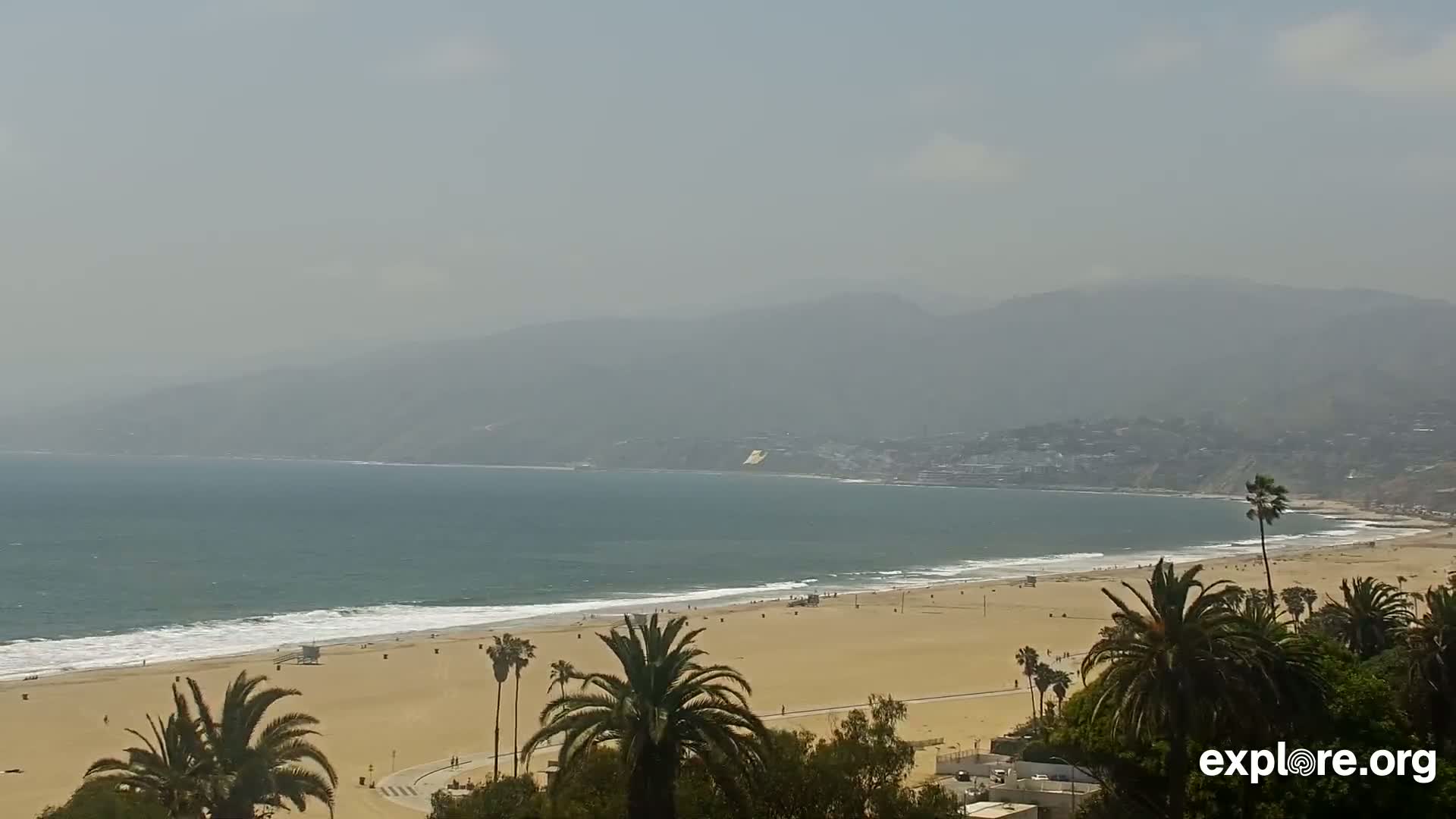A hazy, sunny day shows a long sandy beach with palm trees in the foreground and a calm ocean meeting a distant coastline with mountains under a hazy sky.