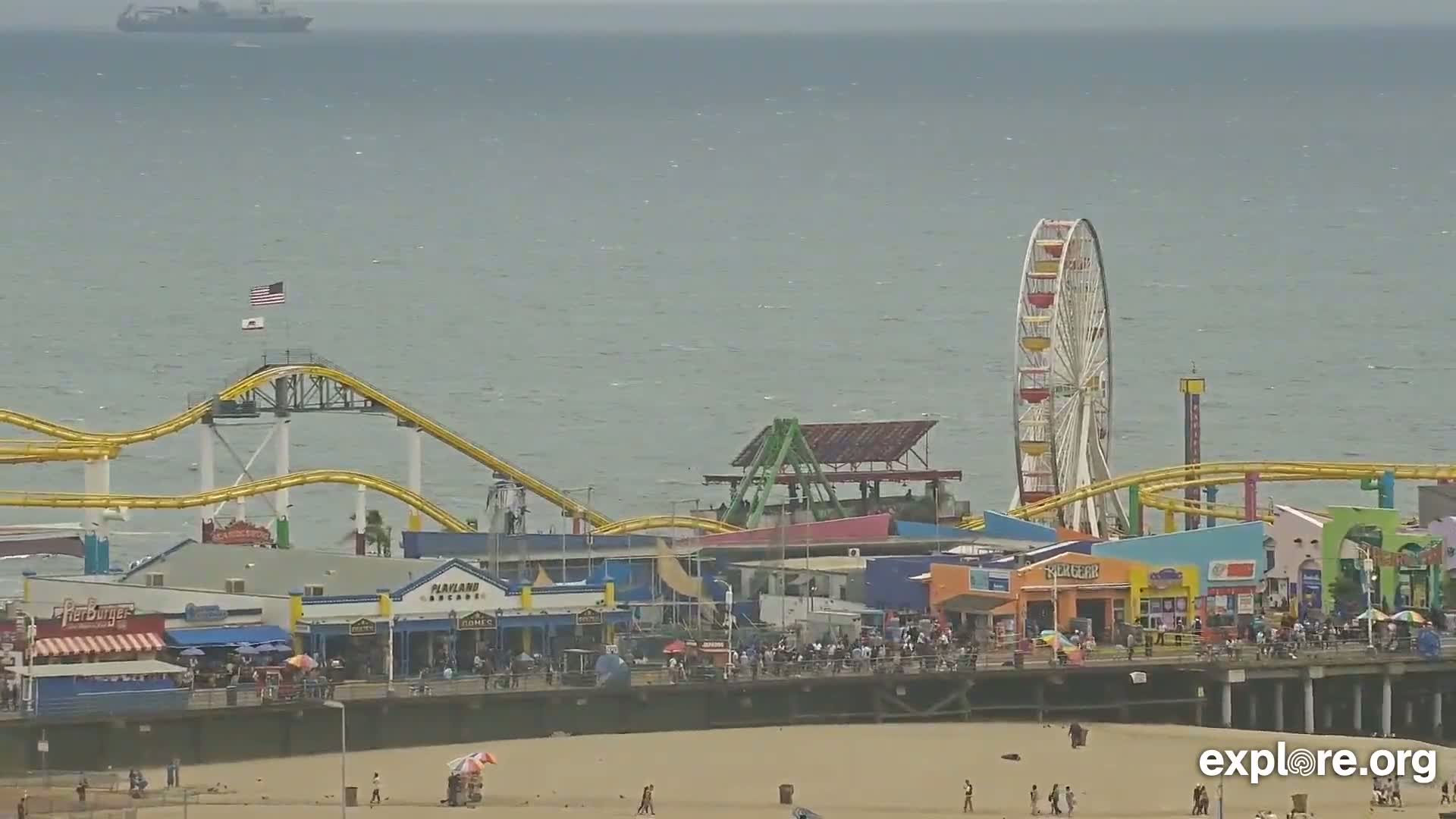 A Ferris wheel and roller coaster are visible on a pier overlooking a beach and ocean under an overcast sky.