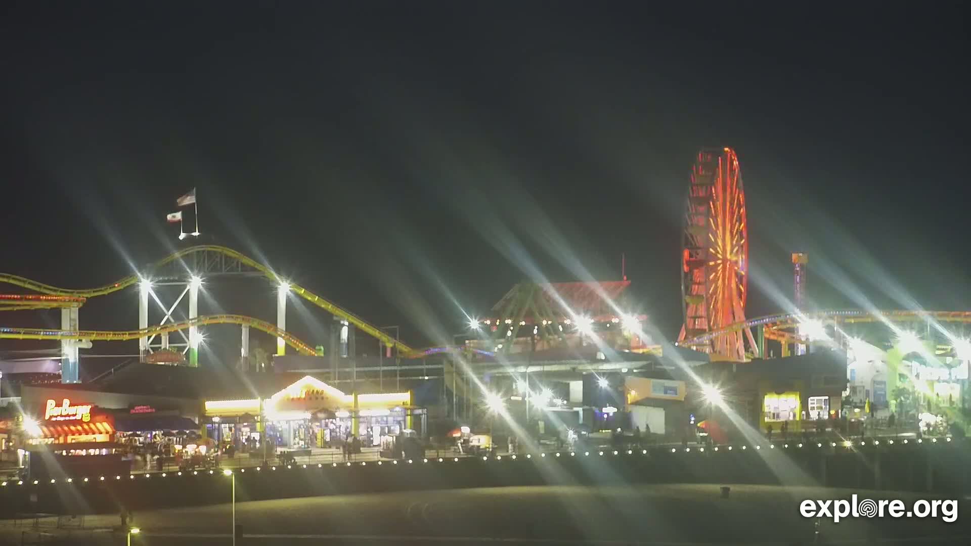 A brightly lit amusement park at night, featuring a roller coaster and Ferris wheel, is seen from a distance.