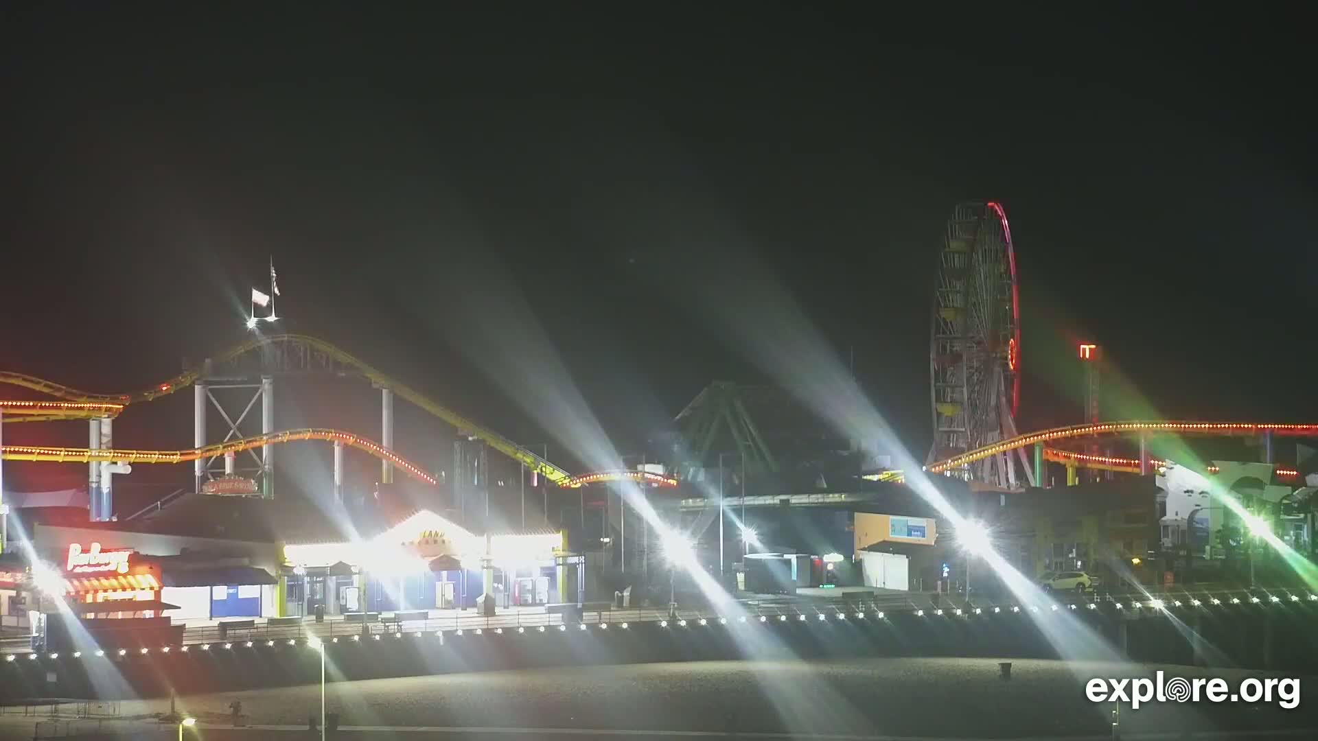 A brightly lit amusement park at night, featuring a roller coaster and Ferris wheel, is illuminated by spotlights against a dark sky.