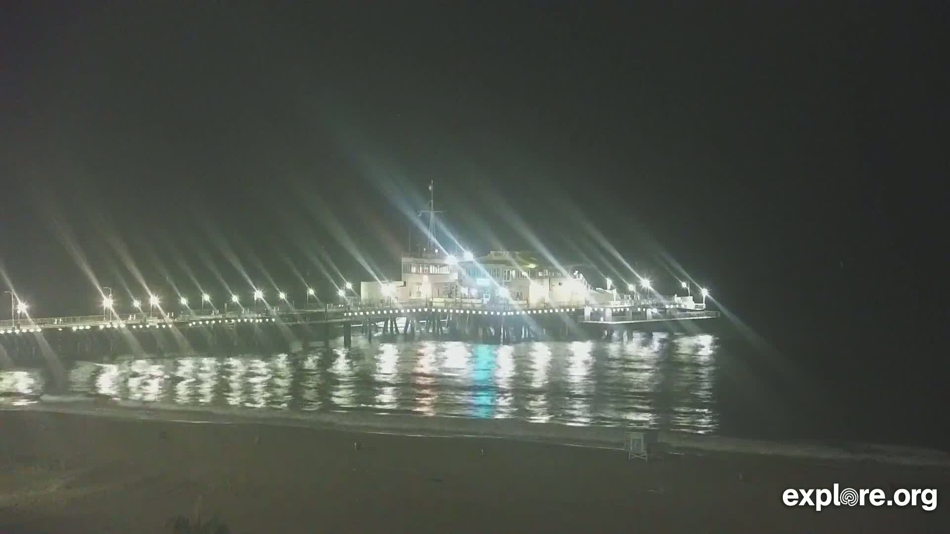 A brightly lit pier extends over a dark, calm ocean at night.