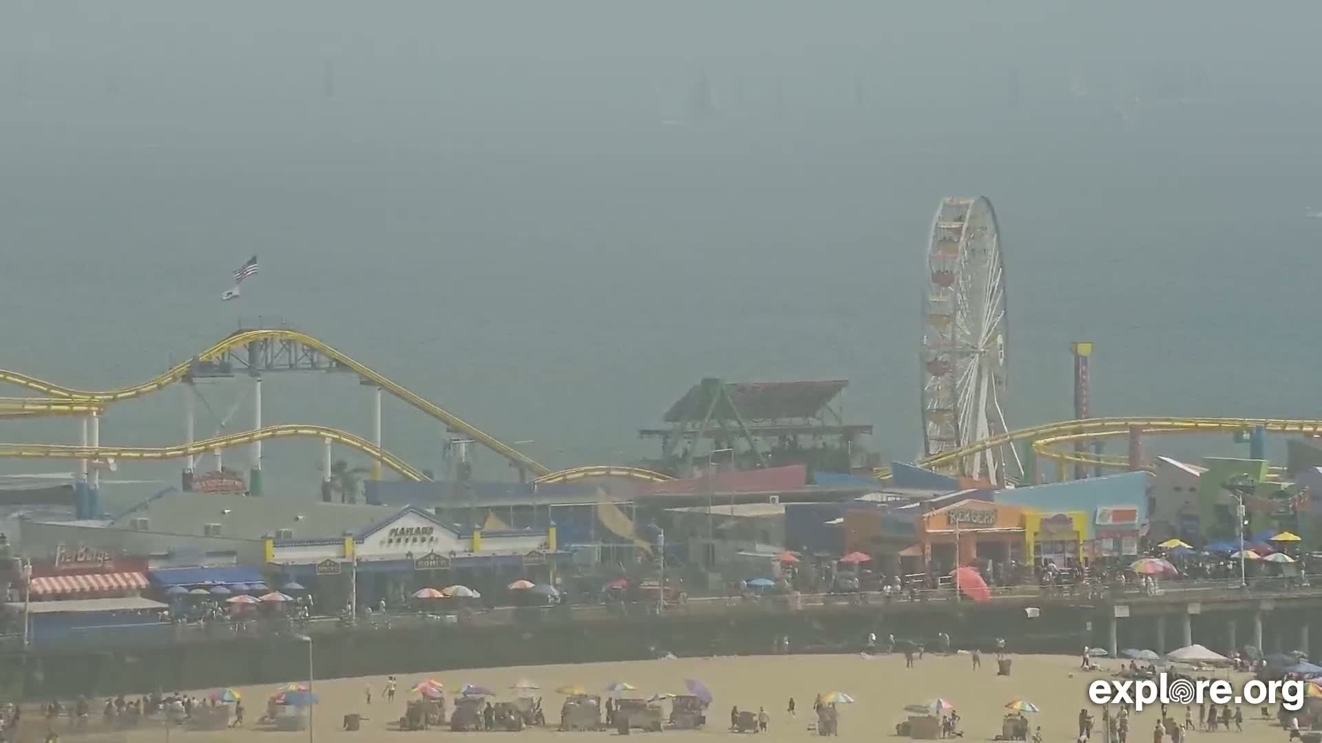 A hazy, sunny day at a beachside amusement park shows a roller coaster, Ferris wheel, and crowds of people on the pier and beach.