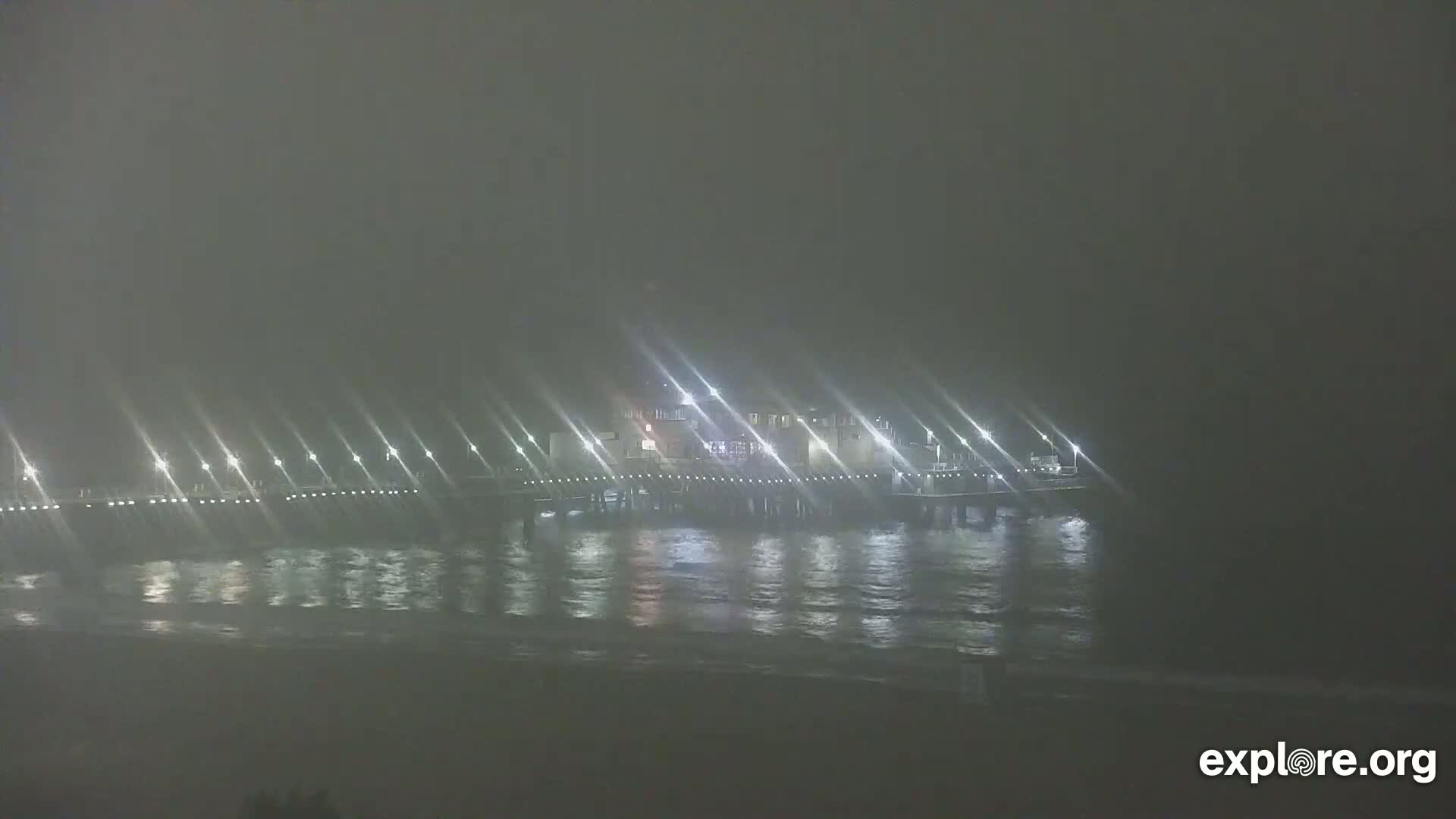 A brightly lit pier extends over a dark, foggy ocean at night.