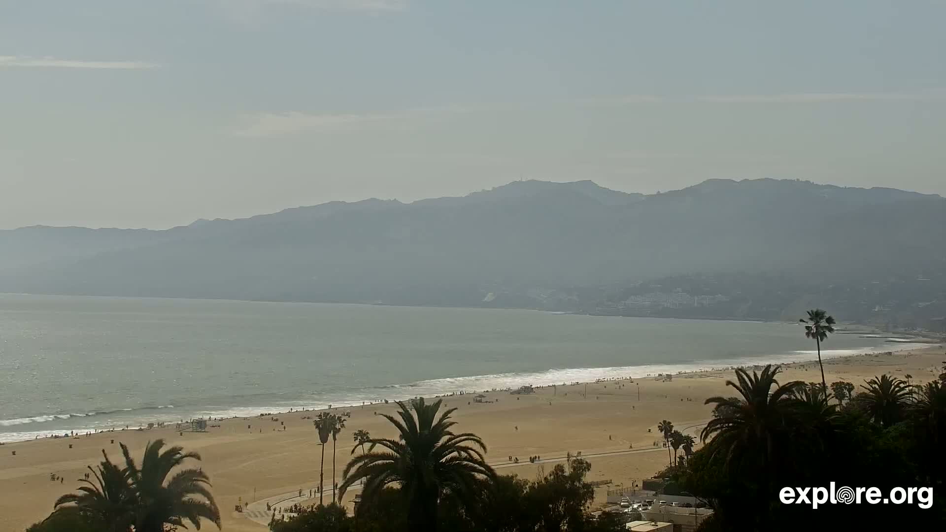 A hazy day reveals a sandy beach with scattered people, palm trees, gentle waves lapping the shore, and mountains in the distance.