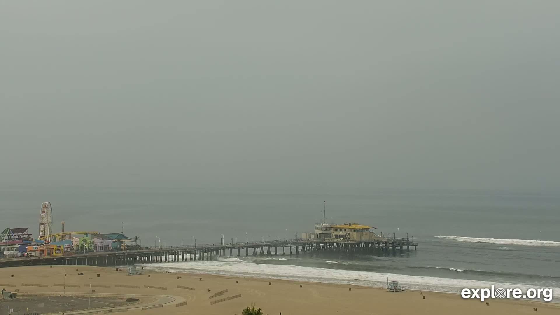 A hazy day reveals a long pier extending over a beach with small waves, and a Ferris wheel visible in the distance.