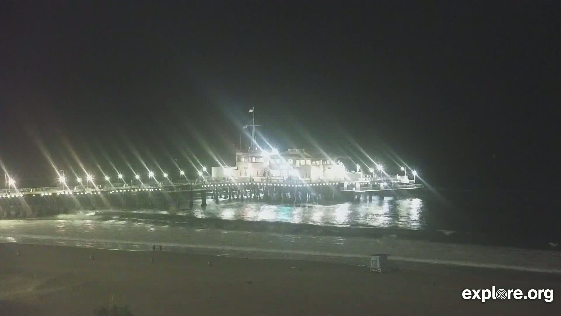 A brightly lit pier extends over dark ocean waves at night, with a sandy beach visible in the foreground.