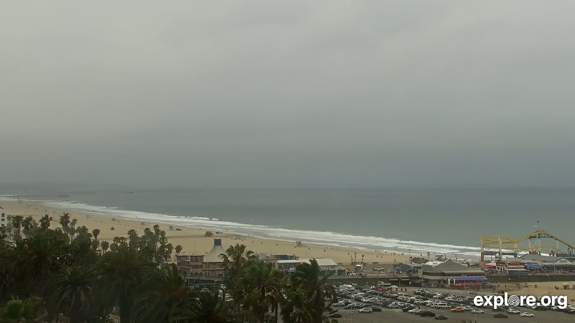 A hazy, overcast day shows a long sandy beach with gentle waves, palm trees, parked cars, and a beachfront amusement park with a roller coaster.