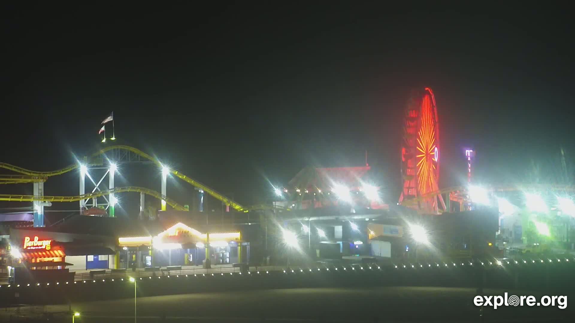 A brightly lit amusement park at night, featuring a roller coaster and Ferris wheel, is slightly obscured by a hazy atmosphere.