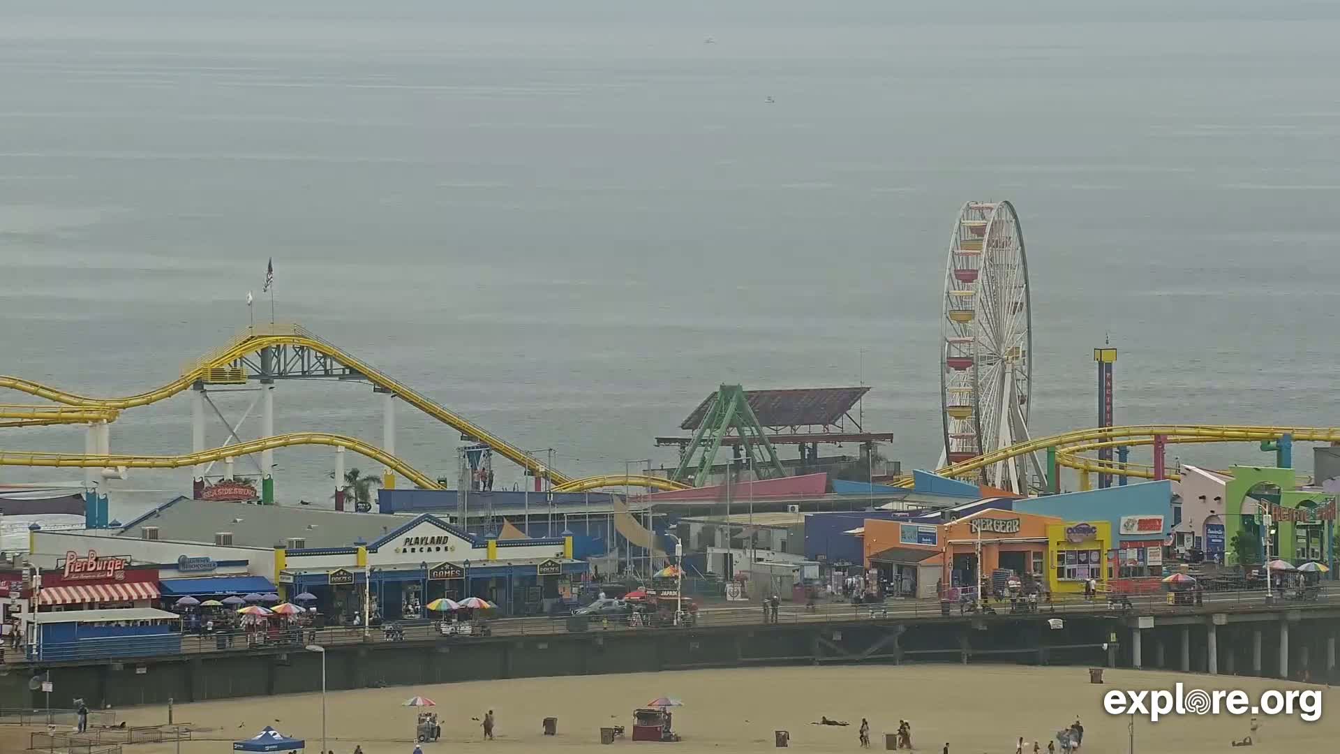 A seaside pier with a roller coaster, Ferris wheel, and various shops and restaurants, on an overcast day.