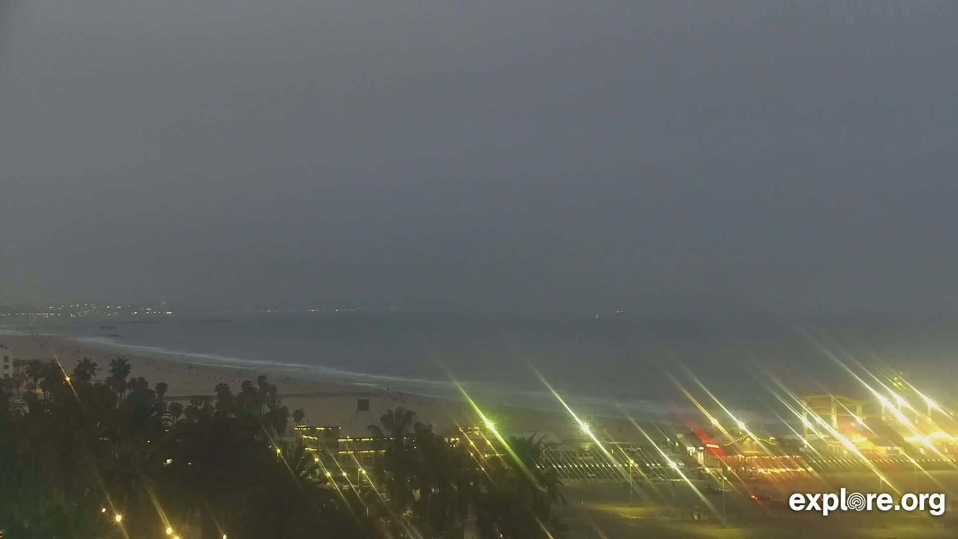 A nighttime beach scene under an overcast sky shows palm trees and lights along a shoreline with the ocean in the background.